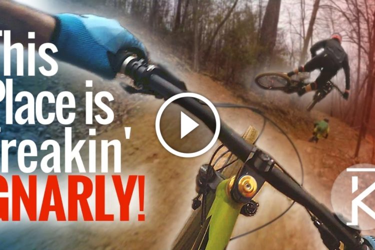 A close-up view of a mountain biker's hand gripping the handlebars on a dirt trail, with another biker performing a jump in the background. The image features the text "This Place is Freakin' GNARLY!" overlaid prominently. The atmosphere conveys excitement and adventure in a challenging outdoor setting.