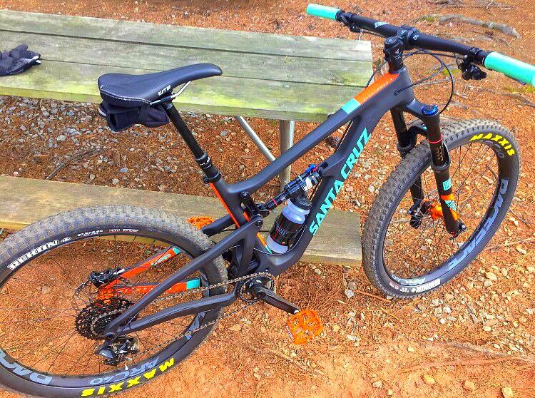Santa Cruz Hightower 29er/27.5: A close-up view of a black and orange mountain bike resting on a dirt surface near a wooden picnic table. The bike features large tires, a prominent suspension system, and vibrant turquoise handlebars. A black seat and a water bottle are visible on the frame.