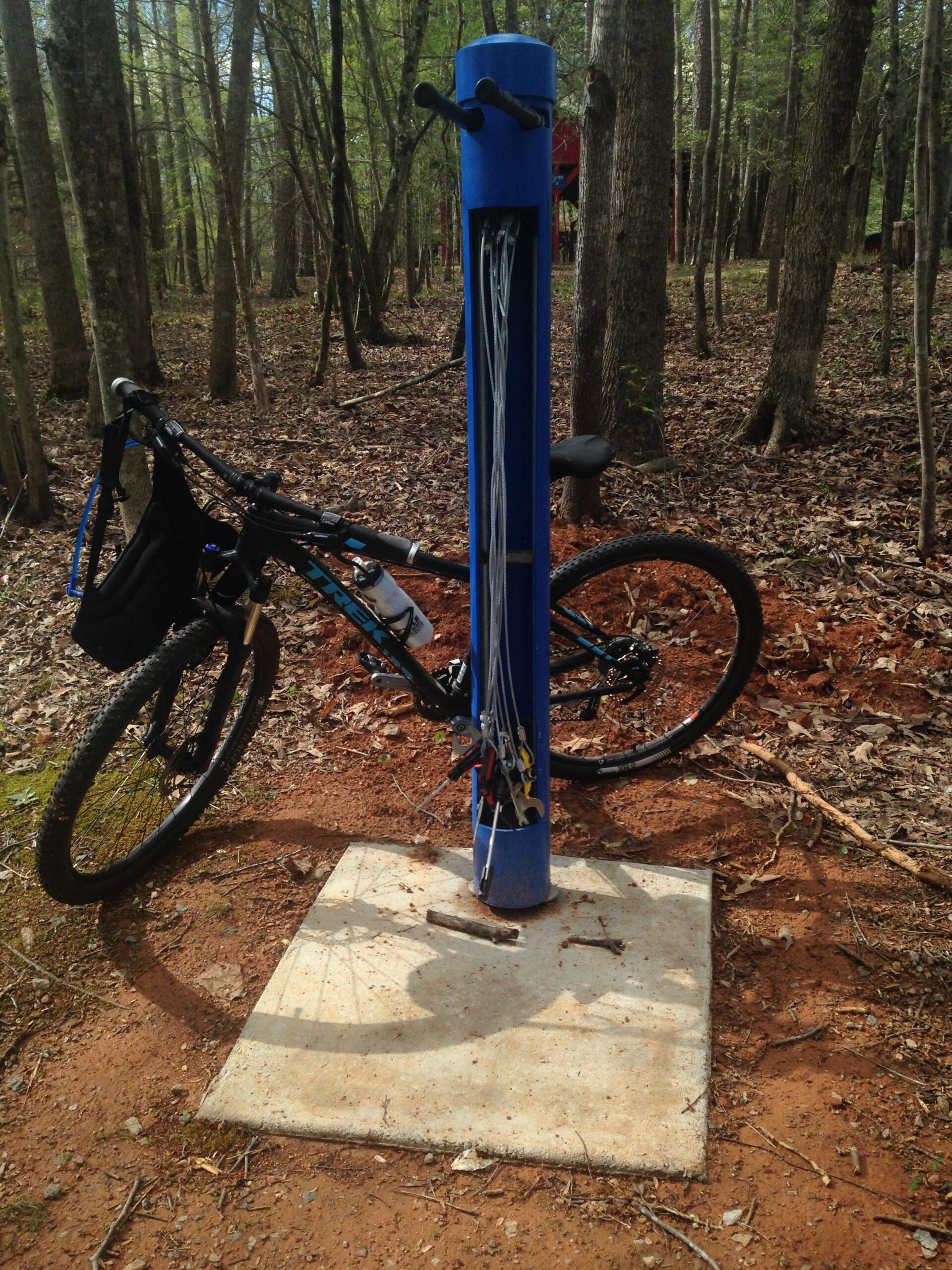Trek X-Caliber 7: A mountain bike parked next to a blue bike repair station in a wooded area, with tools and cables organized on the station. The ground is covered in leaves and dirt, and trees surround the scene.