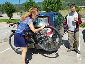 Specialized JETT 29: A person loading a bicycle into the back of a car while another person stands nearby, holding a drink. The setting is an outdoor parking area with trees in the background and a clear blue sky.