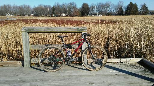 Northrock Northrock XC27: A mountain bike resting on a wooden platform with a railing, set against a backdrop of tall grass and a clear blue sky. The scene captures the bike's muddy tires, suggesting recent use on rugged terrain.