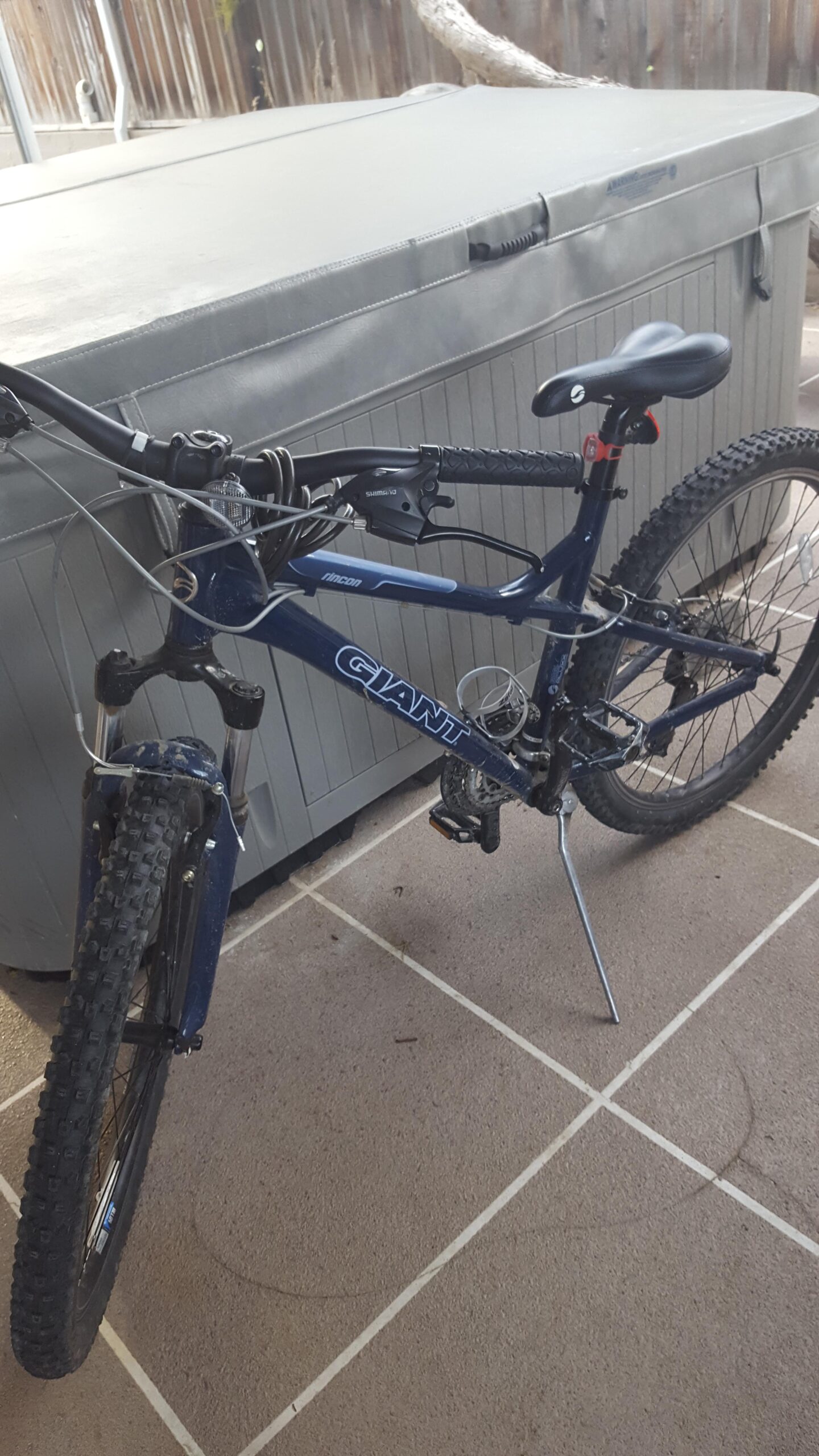 Giant Rincon: A blue Giant Rincon mountain bike is positioned on a tiled patio beside a hot tub. The bike features thick tires, front suspension, and a visible gear system, with the kickstand deployed. The background shows a wooden fence and tree branches.