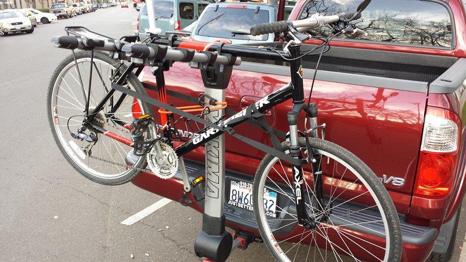 Gary Fisher Tassajara: A black bicycle secured on a bike rack attached to the back of a red pickup truck, parked on a street. The bicycle features racing handlebars, gear system, and is held in place with straps. Surrounding vehicles and a tree can be seen in the background.