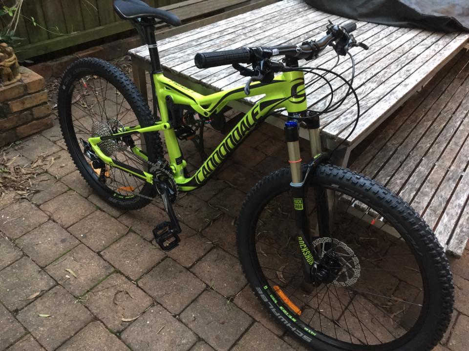 Cannondale Habit: A bright green Cannondale mountain bike positioned on a paved outdoor surface next to a wooden table. The bike features thick tires, front suspension, and a gear shifter, showcasing its design for rugged terrain. There are some outdoor leaves scattered nearby.