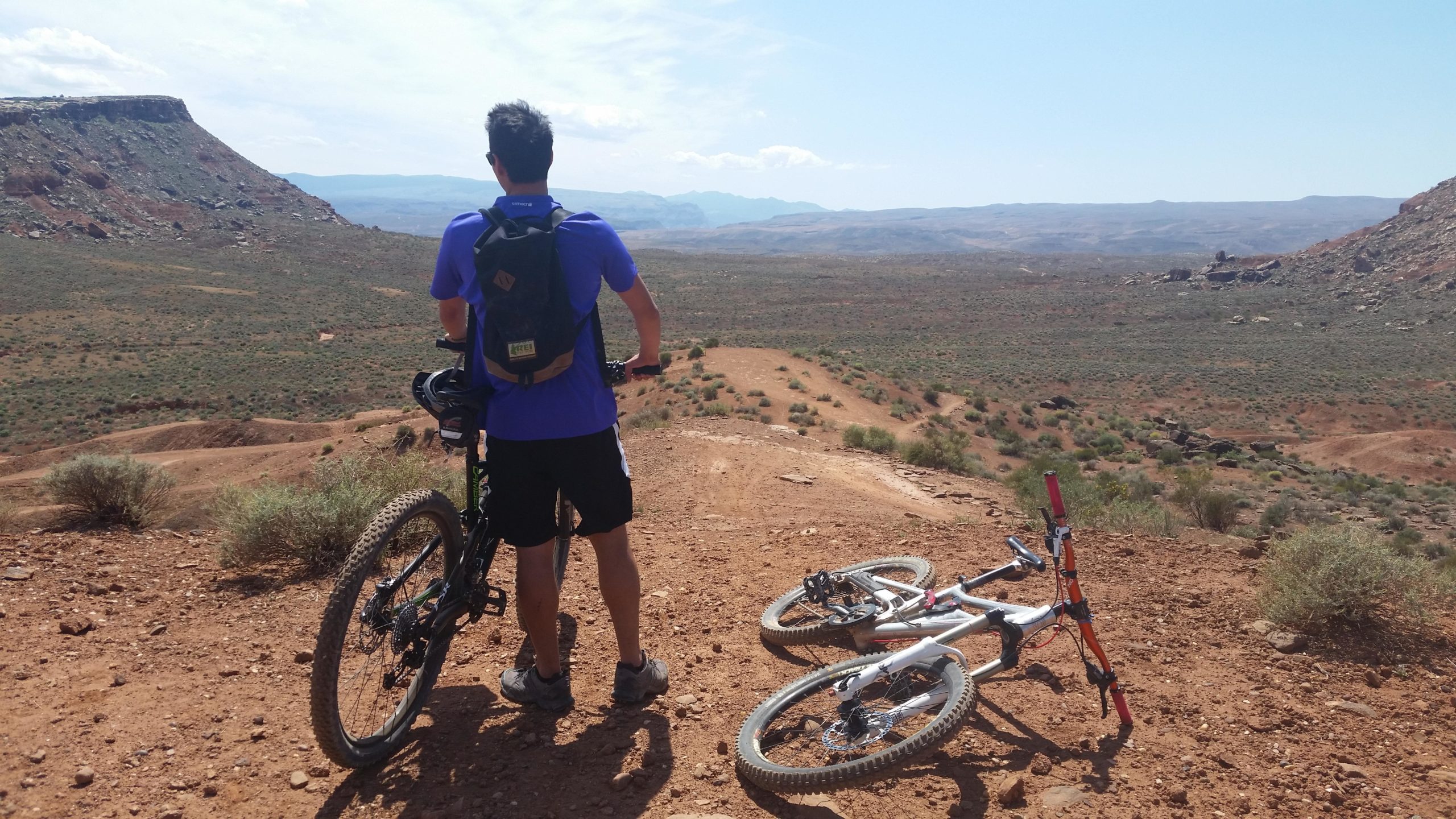 A person in a blue shirt and shorts stands on a dirt trail overlooking a vast, arid landscape. They are holding a mountain bike, while another mountain bike lies on its side nearby. The scene features rocky hills and sparse vegetation under a clear sky. Bearclaw Poppy mountain bike trail.
