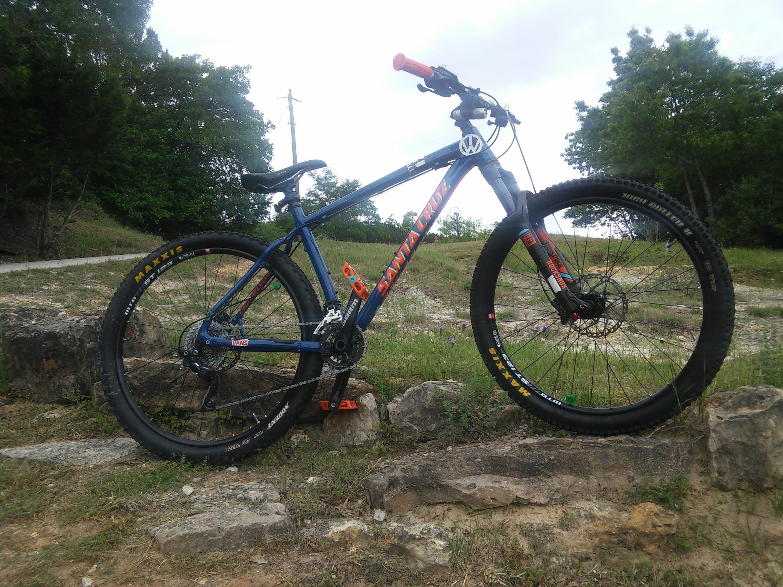 Santa Cruz Chameleon: A mountain bike positioned on a rocky surface, set against a backdrop of green grass and trees. The bike features a blue frame with red accents and is equipped with Maxxis tires. The scene is outdoors, under a partly cloudy sky.
