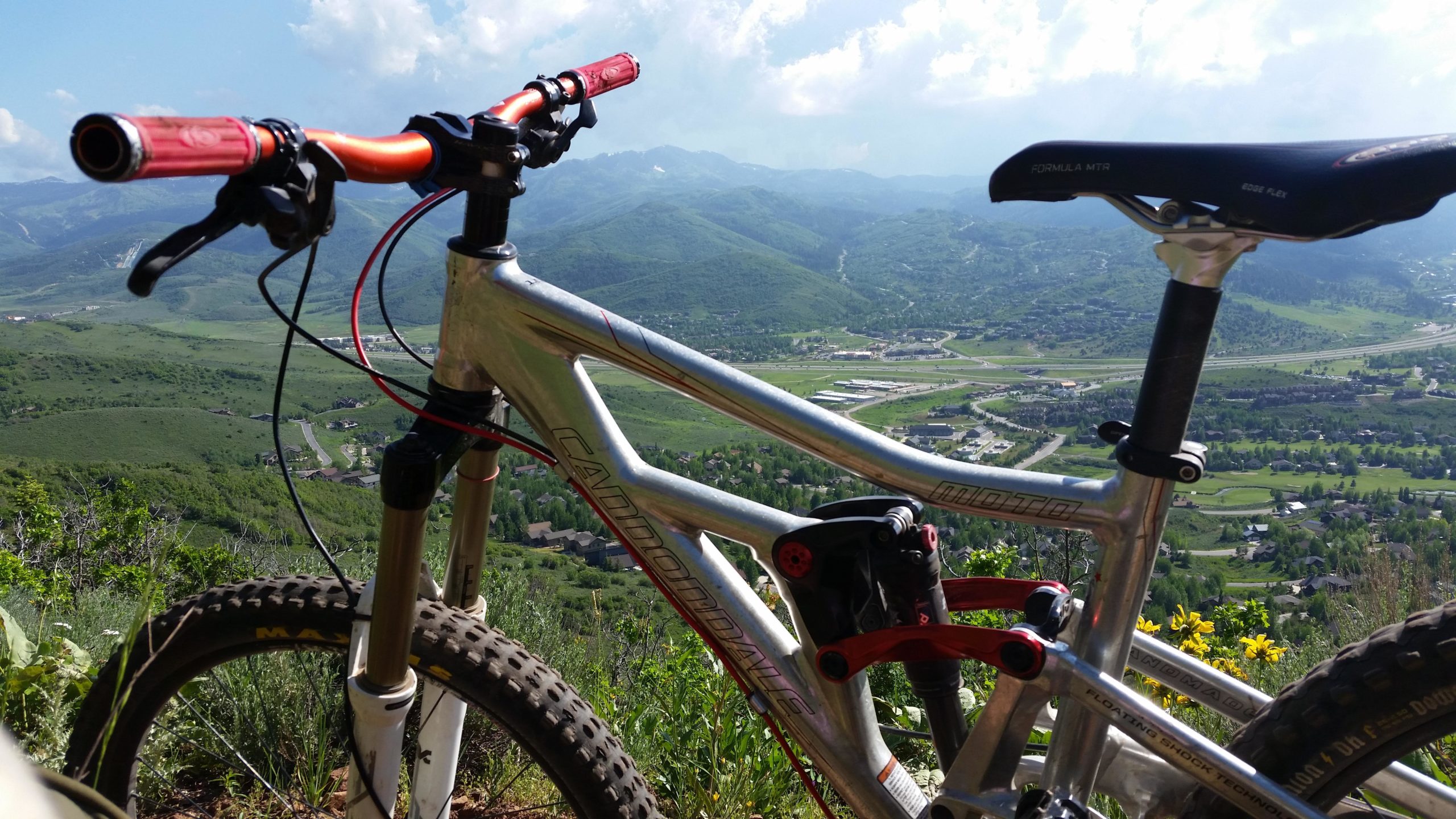 A mountain bike resting on a rocky outcrop, overlooking a lush green valley and distant mountains under a partly cloudy sky. The bike features silver aluminum frame, red handlebars, and thick tires, highlighting a scenic outdoor adventure setting. Glenwild mountain bike trail.