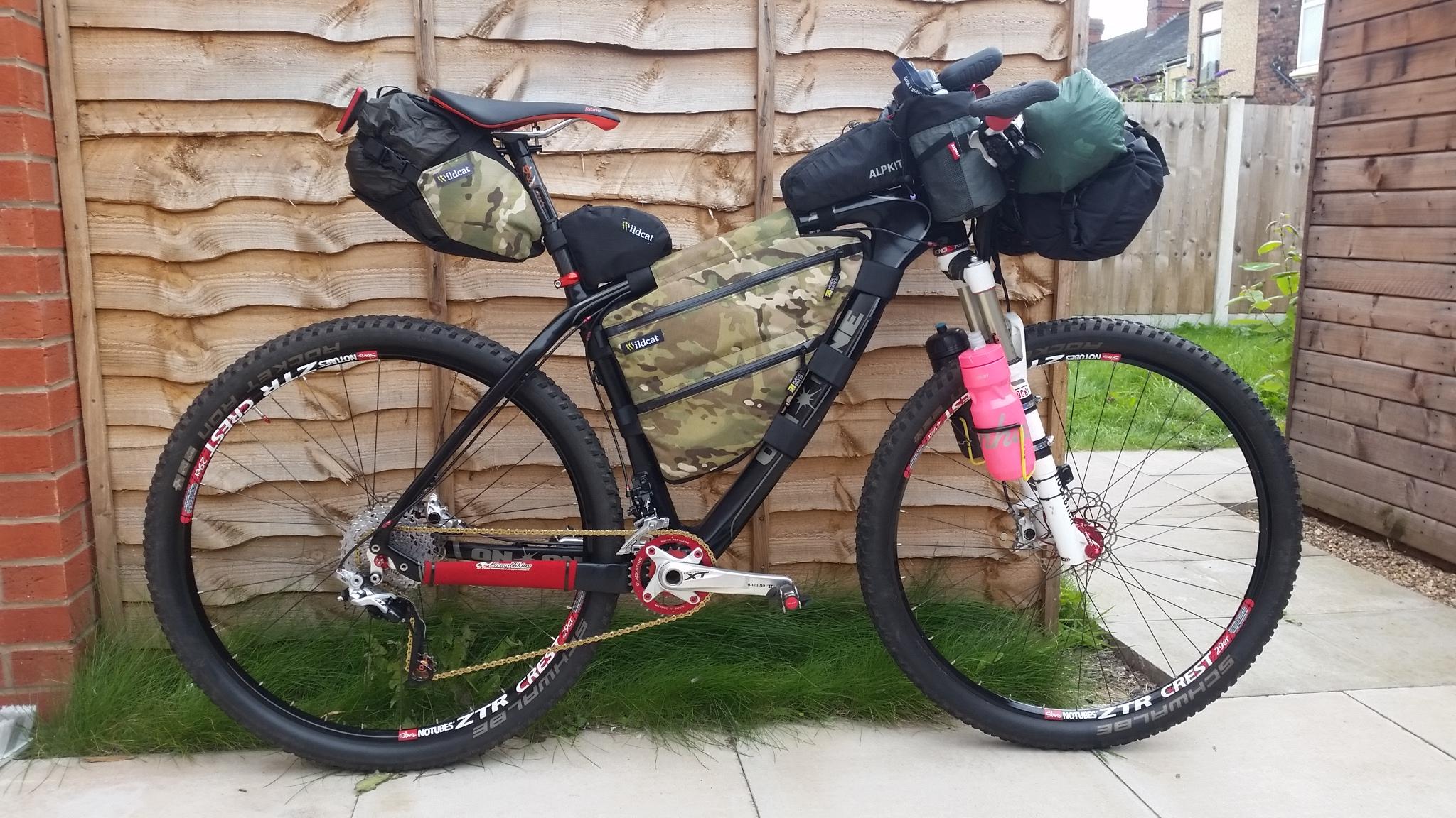 On-One Lurcher: A mountain bike with various bags and equipment attached, featuring a camouflage frame pack and several smaller bags mounted on the handlebars and seat. The bike is positioned against a wooden fence, with green grass in the background and a patio surface beneath. The wheels are thick-tired, suitable for rough terrain, and the bike showcases components designed for durability and performance.
