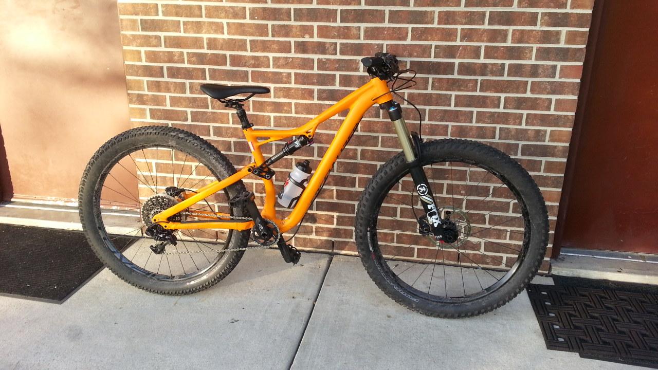 Specialized Stumpjumper FSR 6Fattie: A vibrant orange mountain bike with a sleek frame and thick tires is parked against a brown brick wall. It features a black front suspension fork, a water bottle holder, and an overall rugged design suitable for off-road riding. The area is paved, with a black mat visible on the ground nearby.