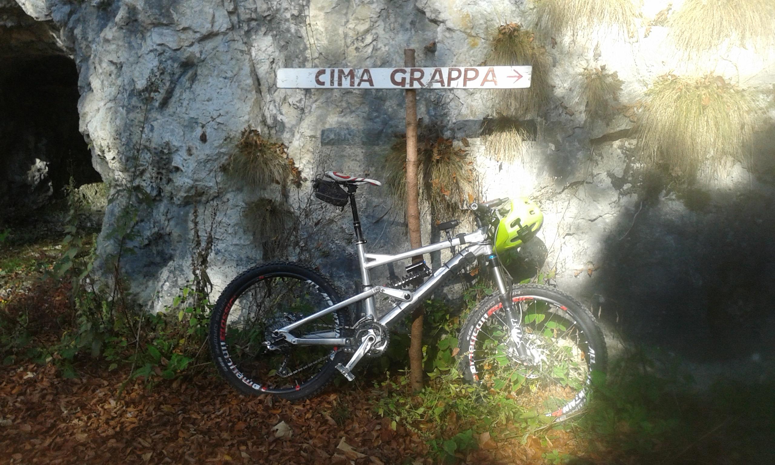 Liteville 301: A mountain bike leaning against a wooden signpost that reads "Cima Grappa" with an arrow pointing in the direction of the trail. The bike features a bright yellow helmet mounted on the handlebars and is situated in a wooded area with rocky terrain and autumn foliage scattered on the ground.