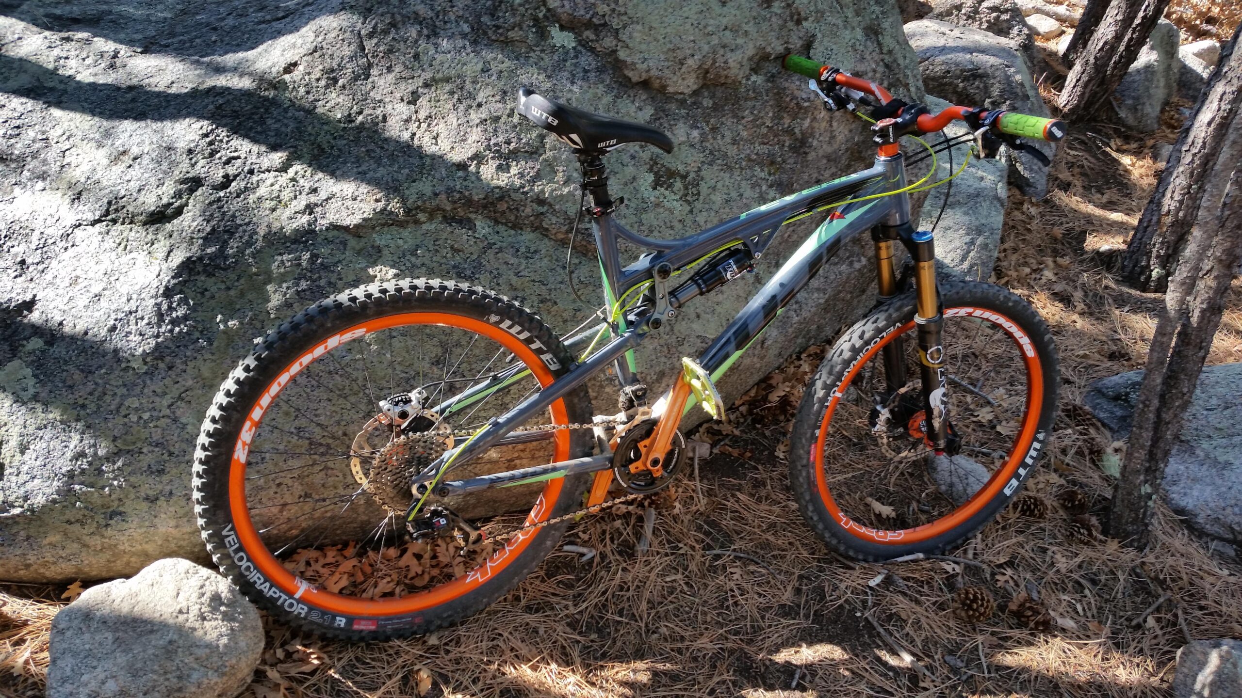 Fuji Reveal 1.0: A mountain bike with a gray frame, orange wheels, and green handlebars rests against a large rock in a forested area. The ground is covered with pine needles and some scattered rocks. Sunlight filters through the trees, casting shadows on the bike.