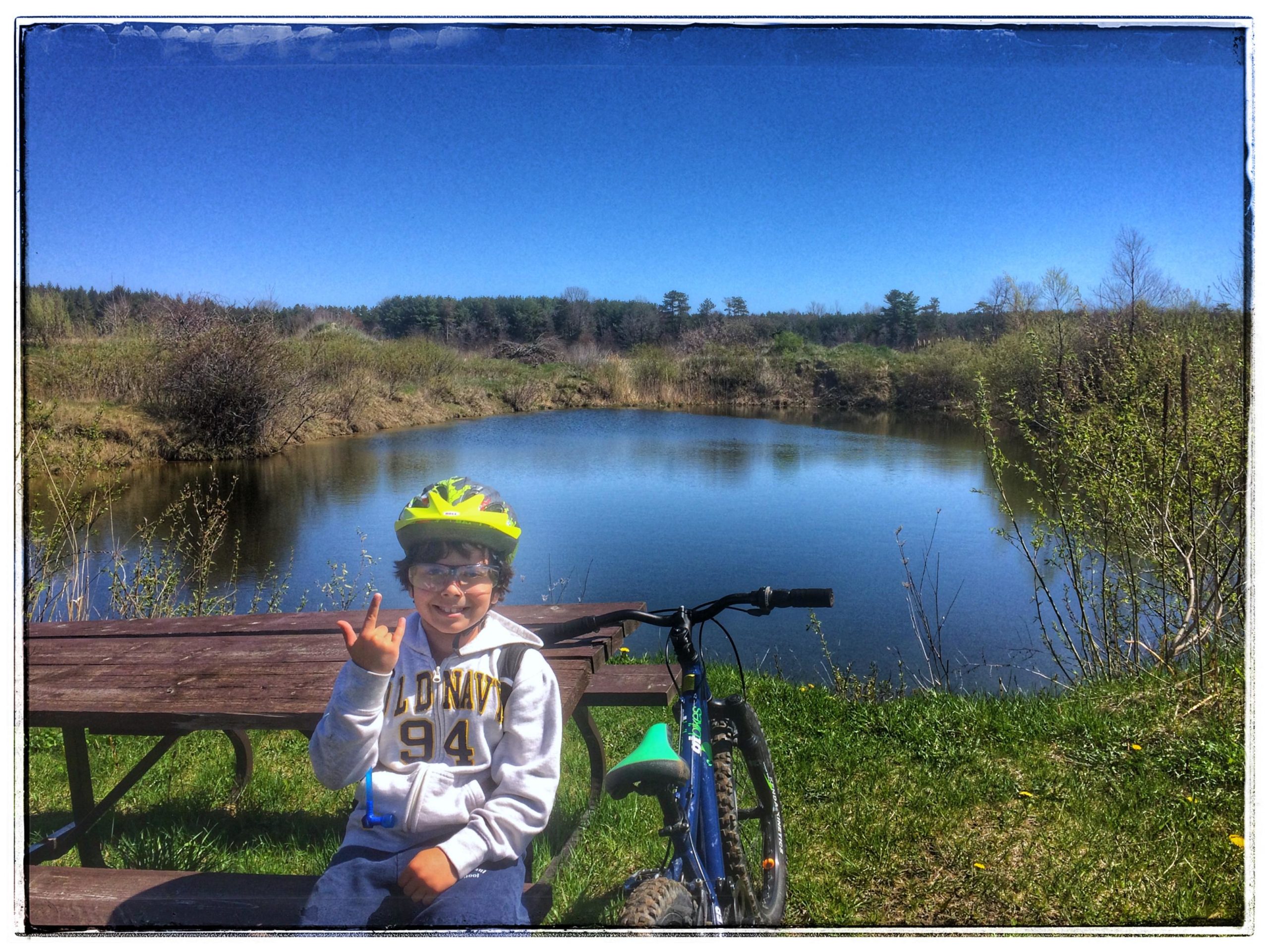 A smiling child wearing a yellow helmet and glasses sits on a picnic bench by a peaceful pond, making a hand gesture. A bicycle rests nearby, surrounded by greenery and trees under a clear blue sky. Turkey Point Provincial Park mountain bike trail.