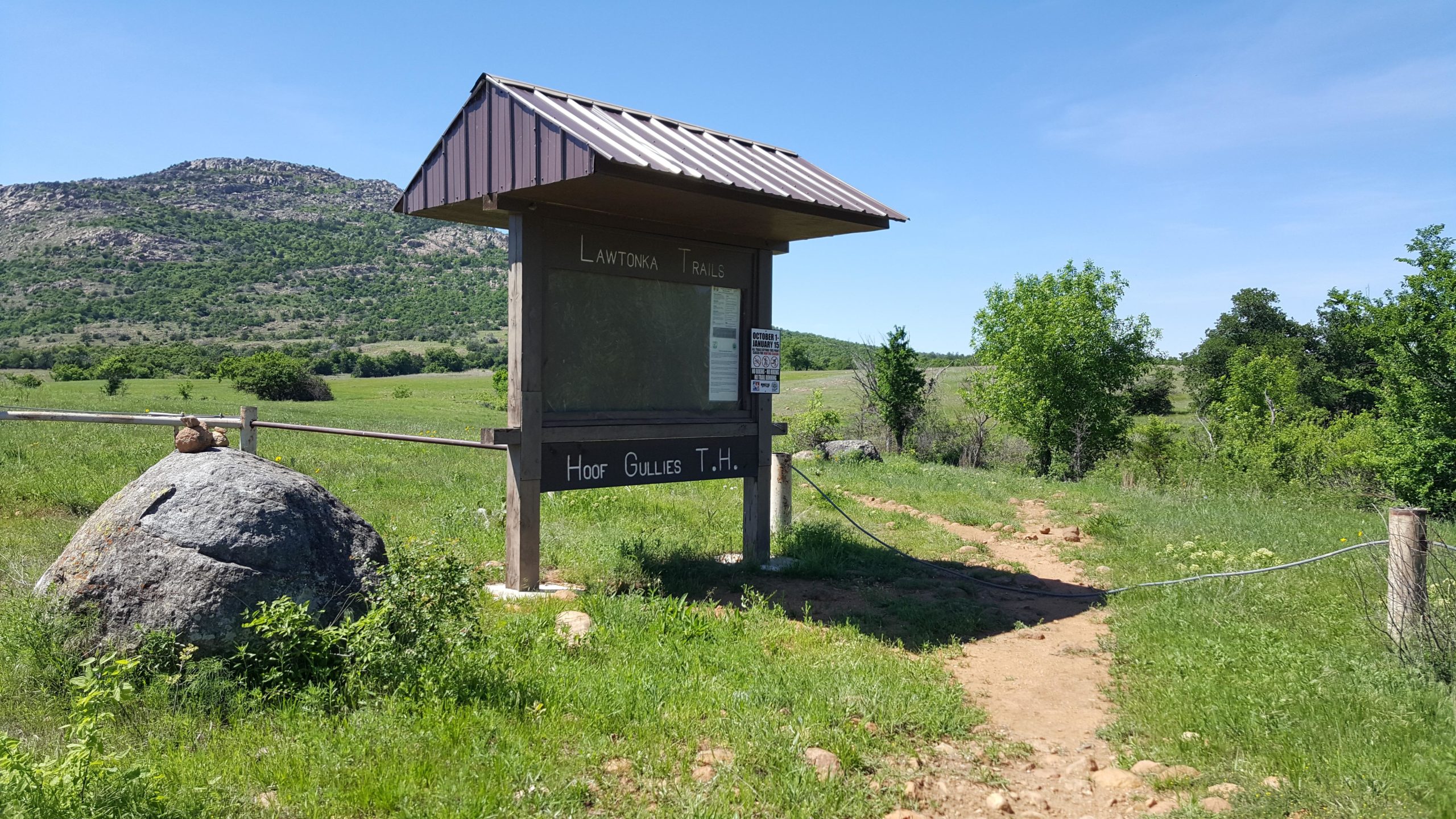 A trailhead sign for Lawtonka Trails labeled "Hoof Gullies T.H." sits near a rocky outcrop, surrounded by green grass and trees. In the background, a rolling hillside is visible under a clear blue sky. A dirt path leads away from the sign into the grassy landscape. Lake Lawtonka Trails mountain bike trail.