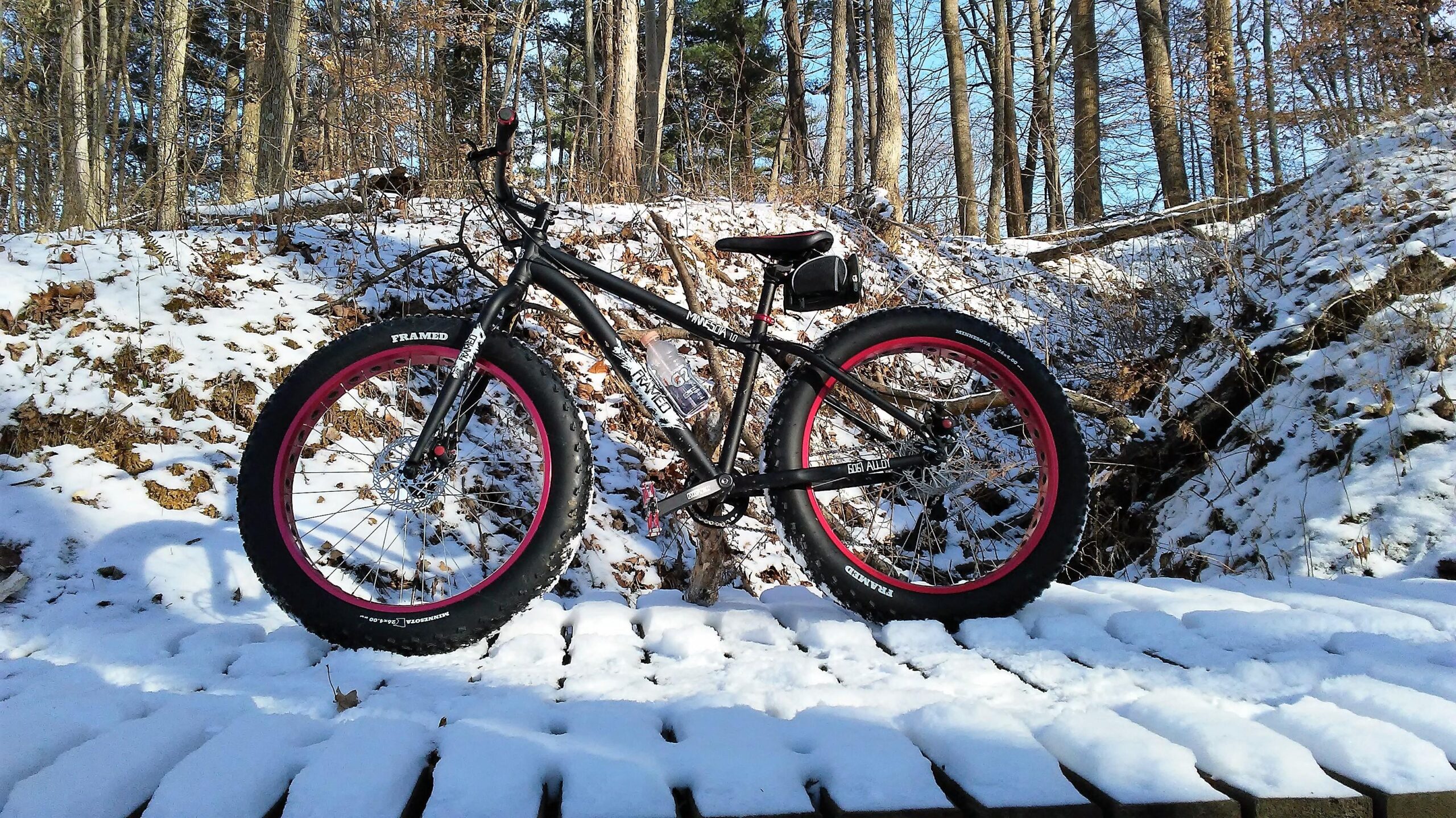Framed Minnesota 1.0: A fat tire bike with black and pink accents is parked on a snowy wooden surface in a winter forest. Background features snow-covered ground and bare trees against a clear blue sky.