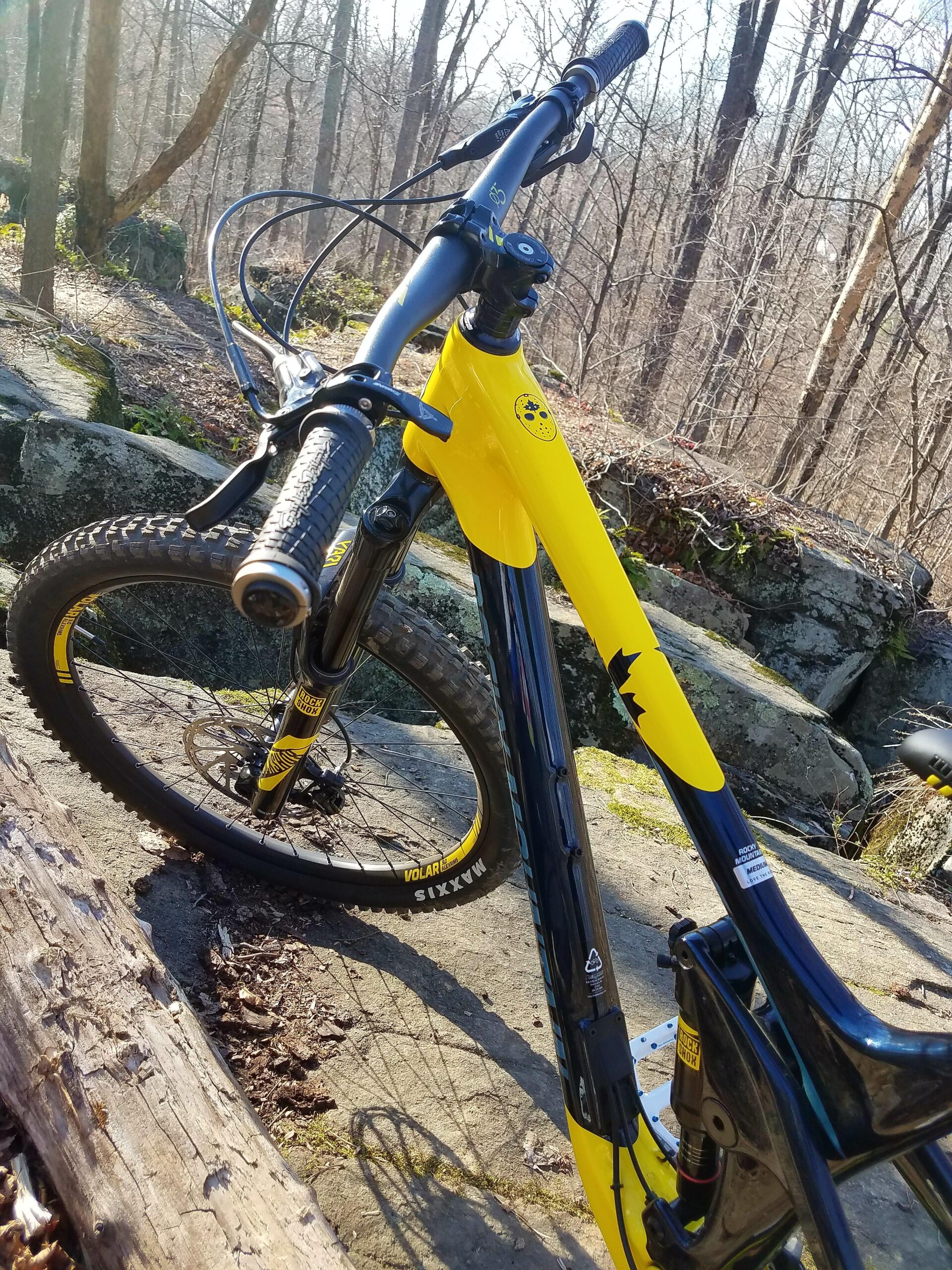 Rocky Mountain Slayer: A close-up view of a mountain bike featuring a bright yellow frame with black accents, positioned on a rocky surface in a forested area. The handlebars and front wheel are prominently visible, with tree branches and foliage in the background, indicating an outdoor setting.