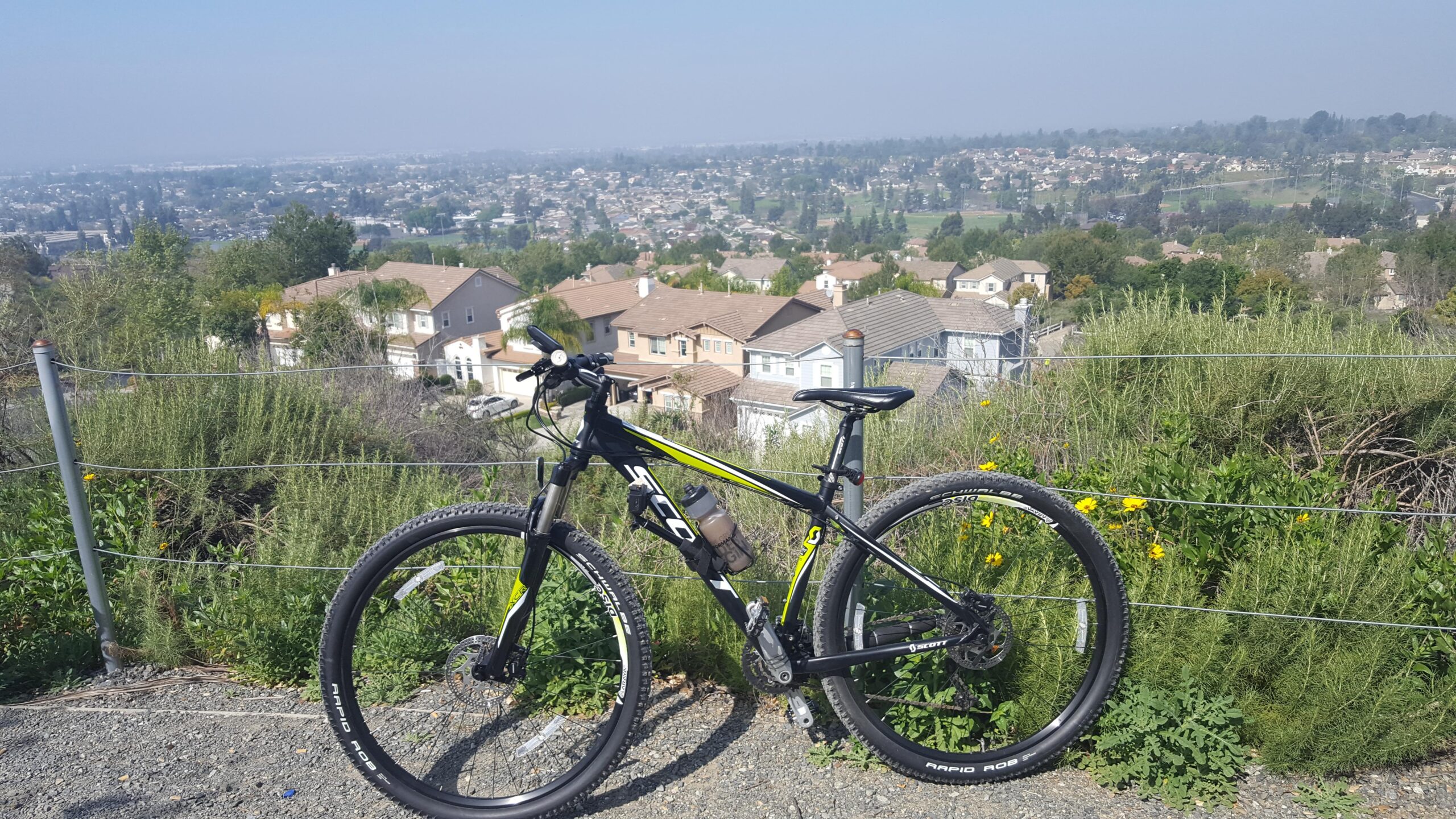 Scott Aspect Trail 29er: A mountain bike resting on gravel with a scenic view of houses and greenery in the background. The landscape features a hazy skyline with trees, homes, and a distant neighborhood. Bright yellow flowers and lush bushes surround the bike, enhancing the outdoor setting.