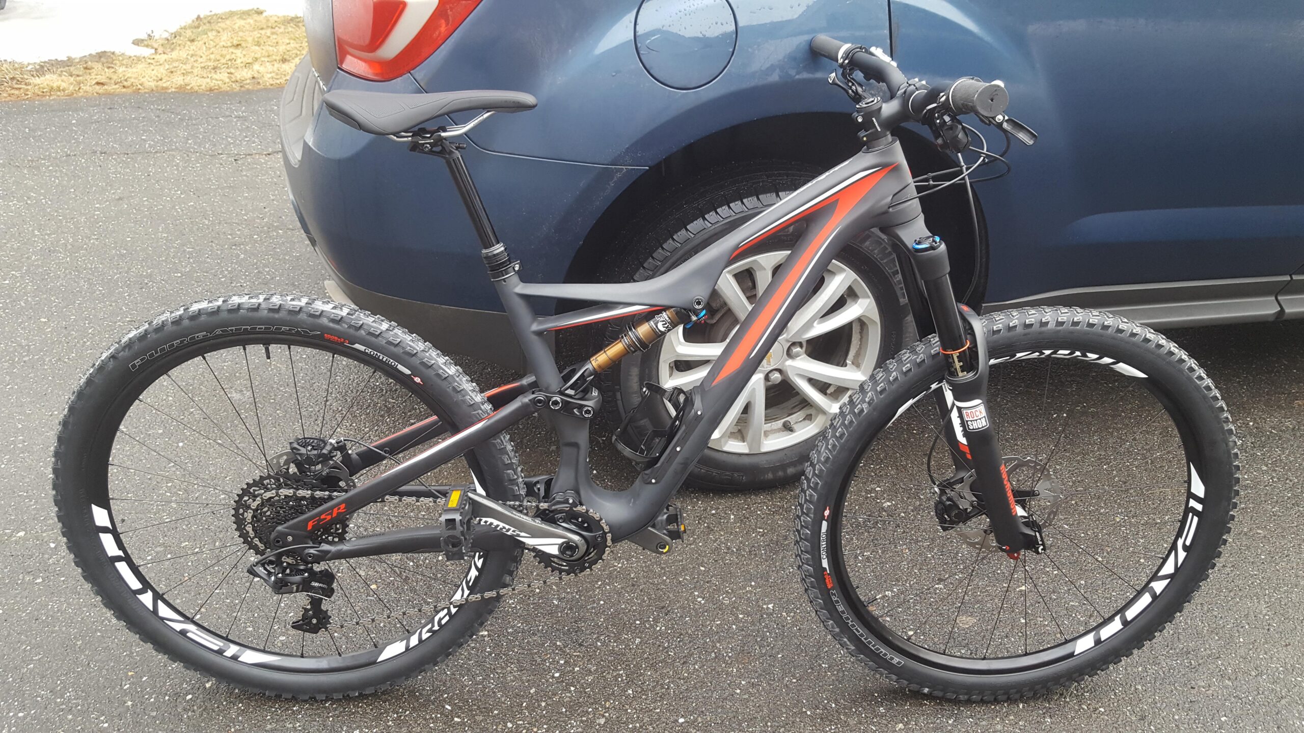 Specialized Stumpjumper FSR Expert Carbon 650B: Mountain bike parked next to a blue car, showcasing its dark frame with red accents, thick tires, and suspension components. The bike is positioned on a paved surface, with the car's rear tire partially visible in the background.