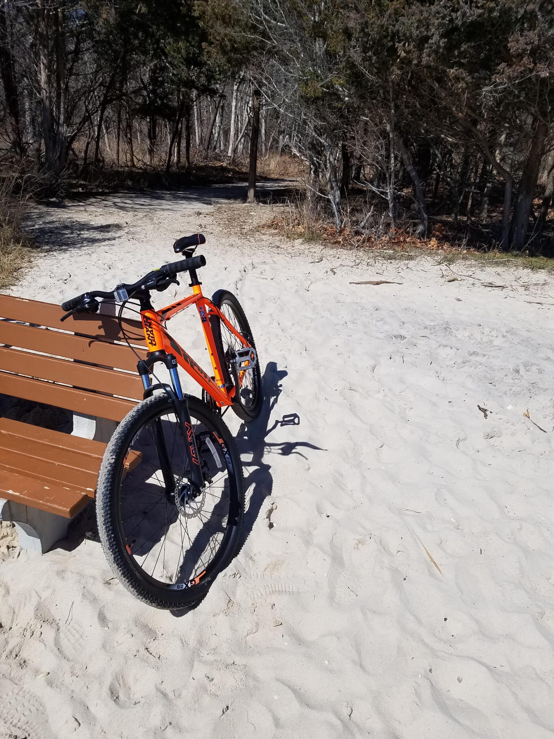 Giant ATX 27.5 2: A vibrant orange mountain bike rests against a wooden bench, situated on sandy ground. In the background, there are patches of trees and a clear path leading into a wooded area, suggesting a natural outdoor setting. The scene conveys a sense of adventure and tranquility.