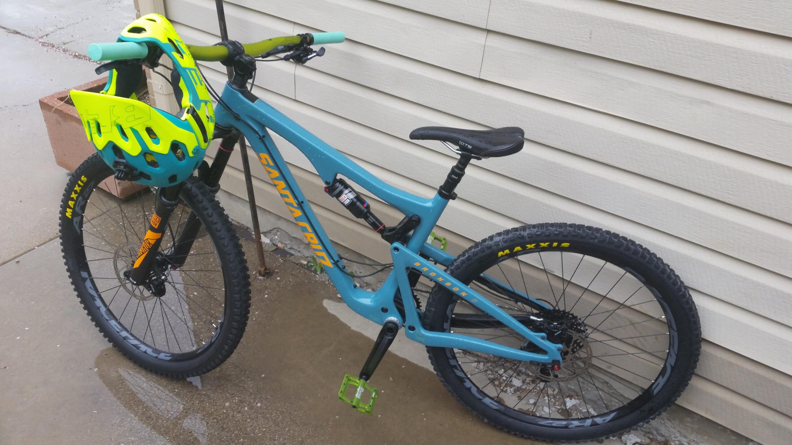 Santa Cruz Bronson: A blue mountain bike with thick tires, a green and yellow helmet resting on the handlebars, and green pedal clips. The bike is positioned next to a building with textured siding, and there are small puddles of water on the ground, suggesting recent rain.
