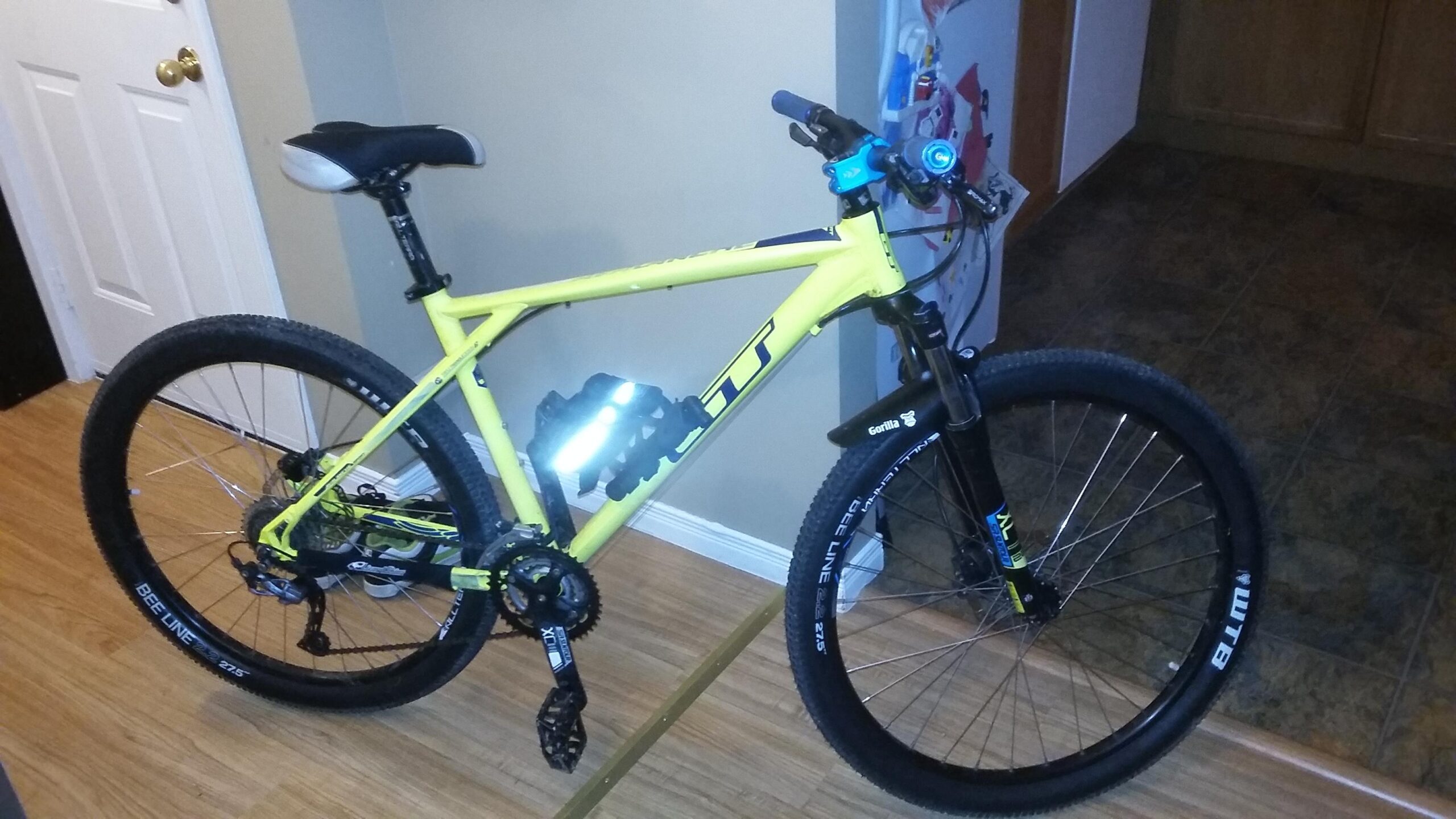 GT Avalanche: A bright yellow mountain bike with black wheels and components, positioned at an angle in a room with wood flooring. The bike features a sleek frame, suspension forks, and accessories such as a water bottle holder and a front light. A door can be seen in the background along with a refrigerator.