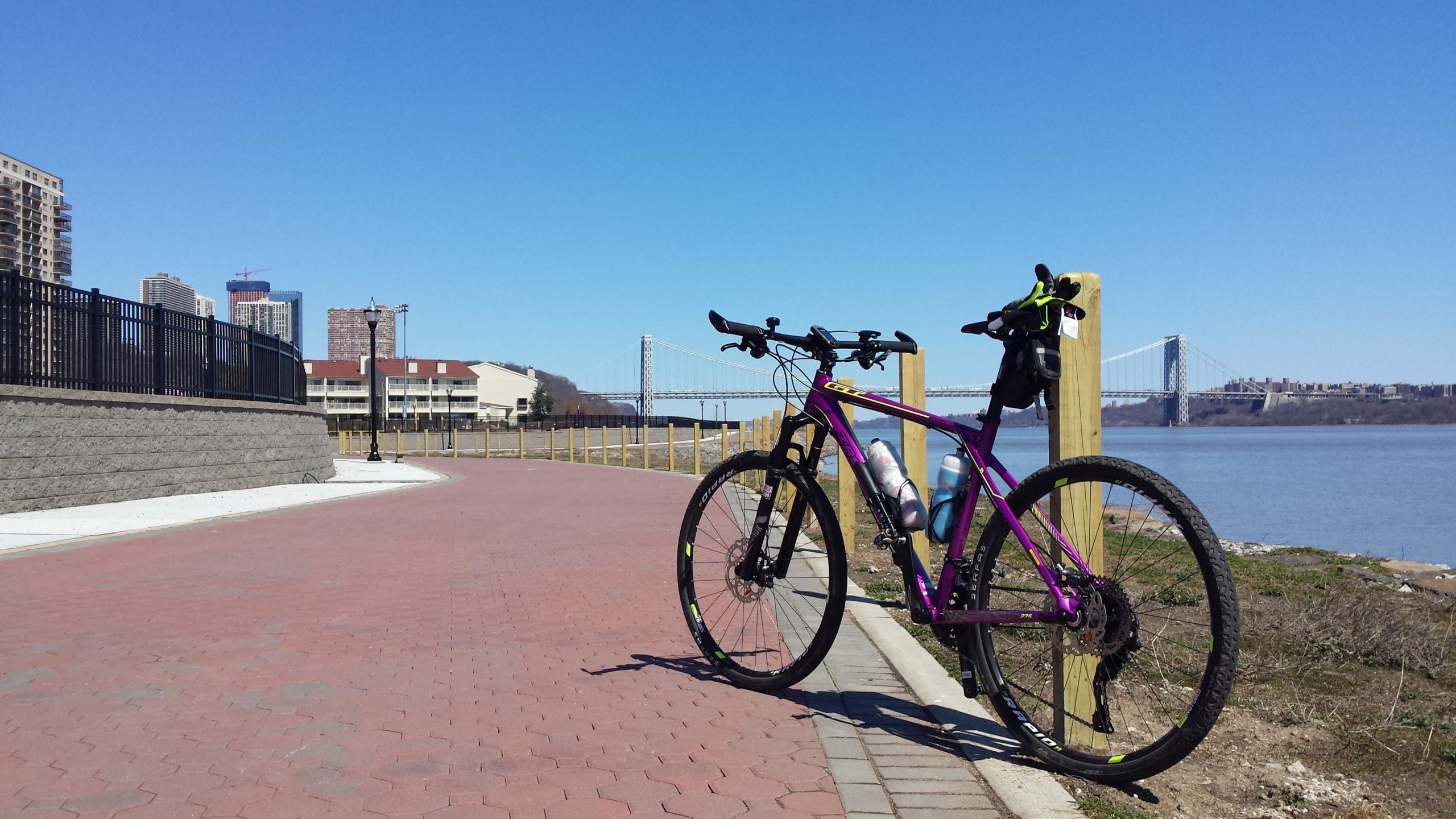A purple bicycle is parked along a paved pathway by a river, with a view of buildings and a bridge in the background. The sky is clear and blue, indicating nice weather. West Street Greenway mountain bike trail.