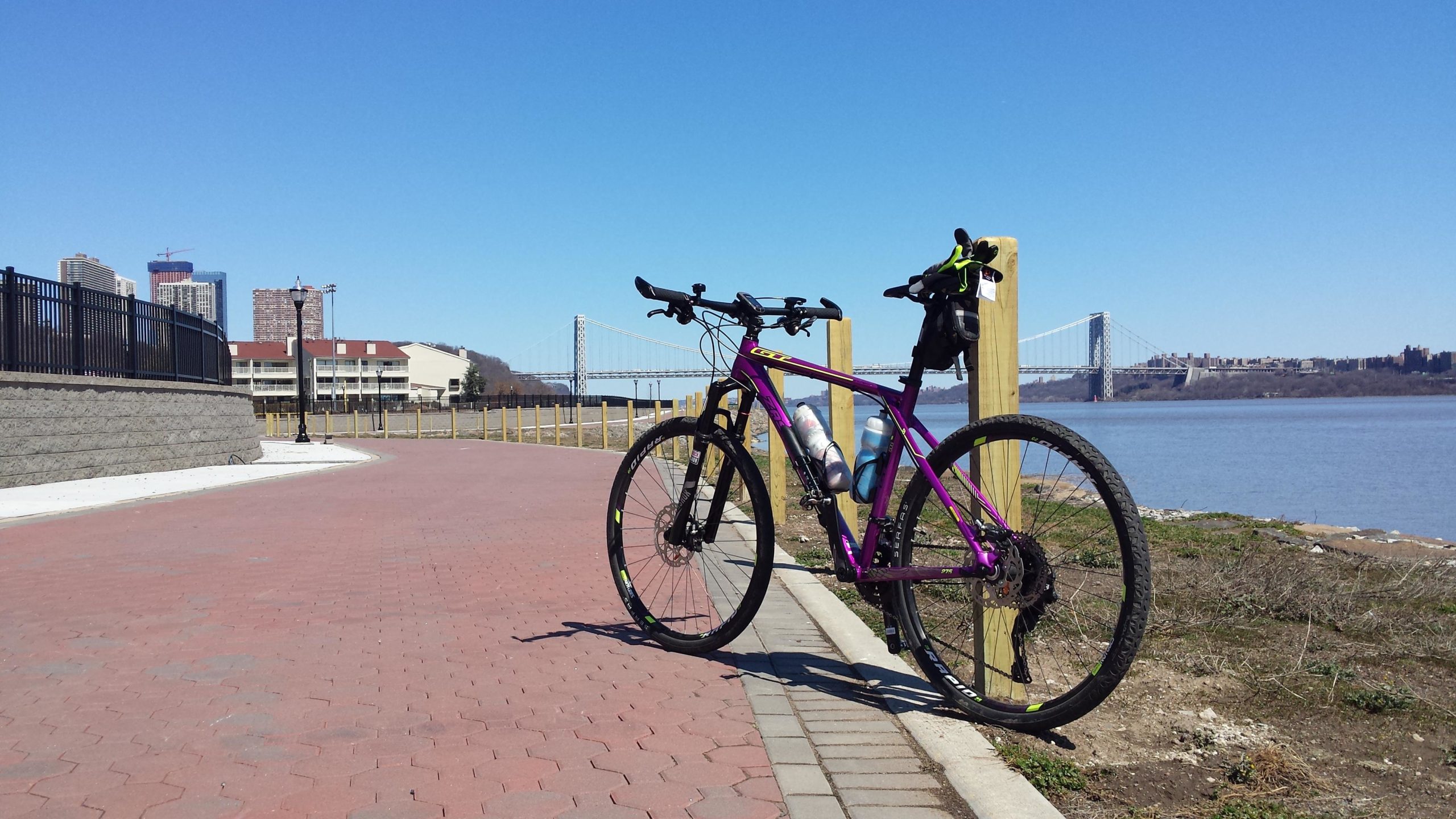 A purple mountain bike stands on a paved path next to a river, with the George Washington Bridge visible in the background. The scene is set on a clear day, featuring a bright blue sky and urban buildings on the left side. The bike is equipped with water bottles and a bag attached to its frame. West Street Greenway mountain bike trail.