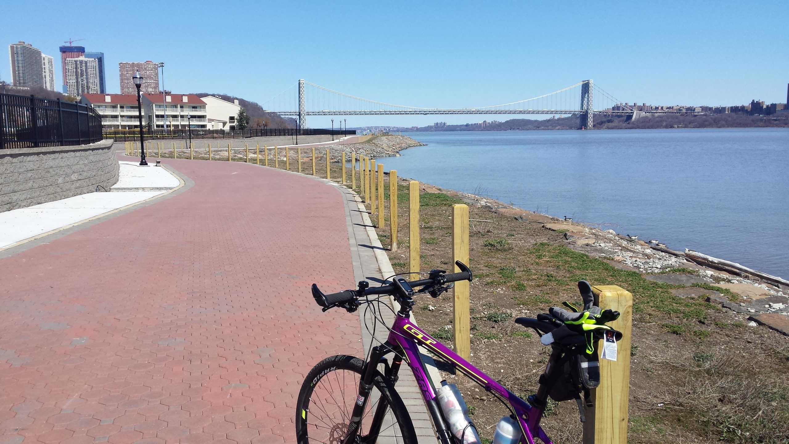 A bike parked along a paved pathway beside a river, with a bridge in the background and city skyline partially visible. The scene is set on a clear day, showcasing blue skies and calm water. West Street Greenway mountain bike trail.