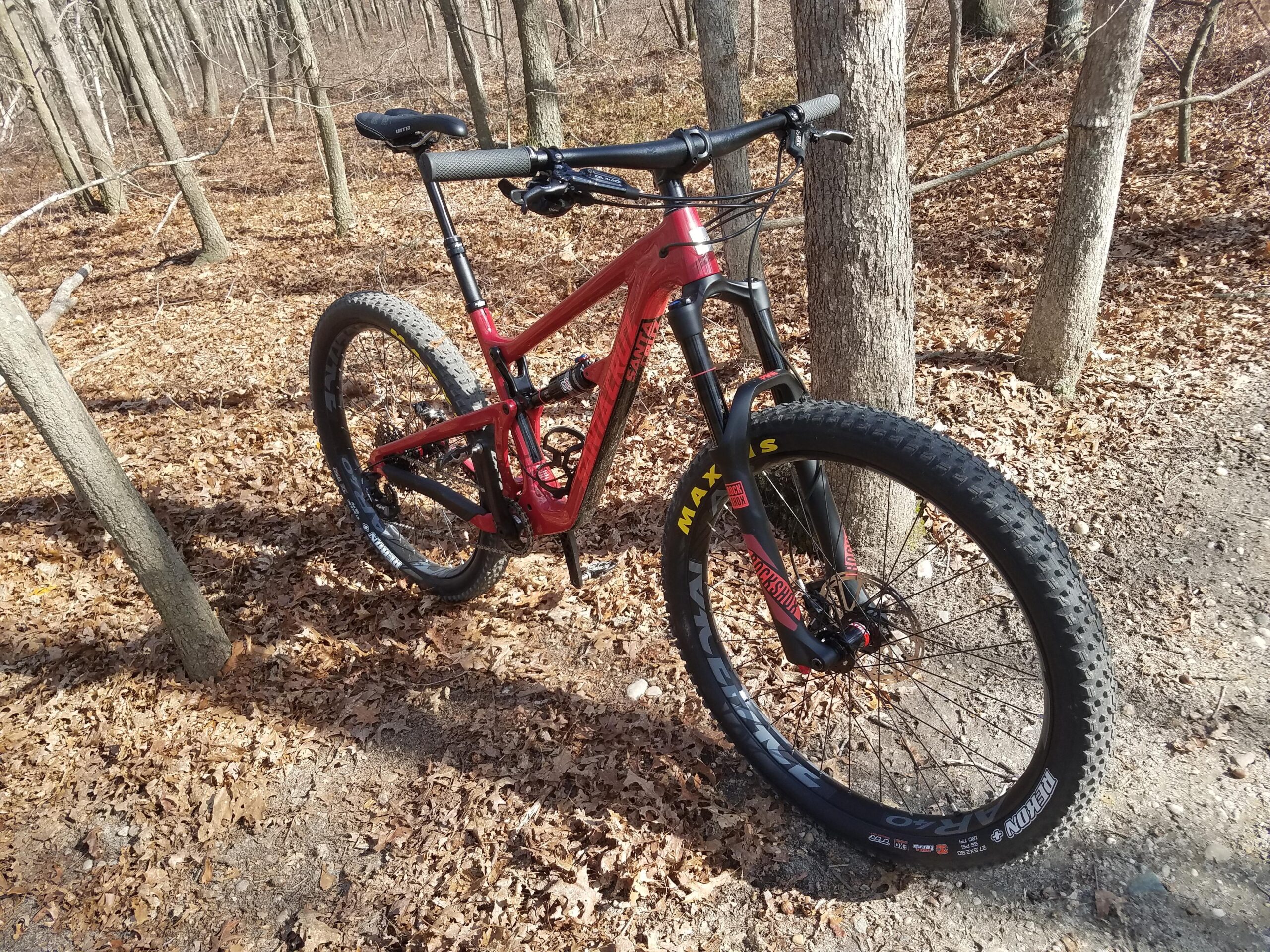 Santa Cruz Hightower 29er/27.5: A red mountain bike parked on a leaf-covered trail in a forest, surrounded by trees. The bike features thick tires and is positioned next to a tree, with additional foliage and a dirt path visible in the background.