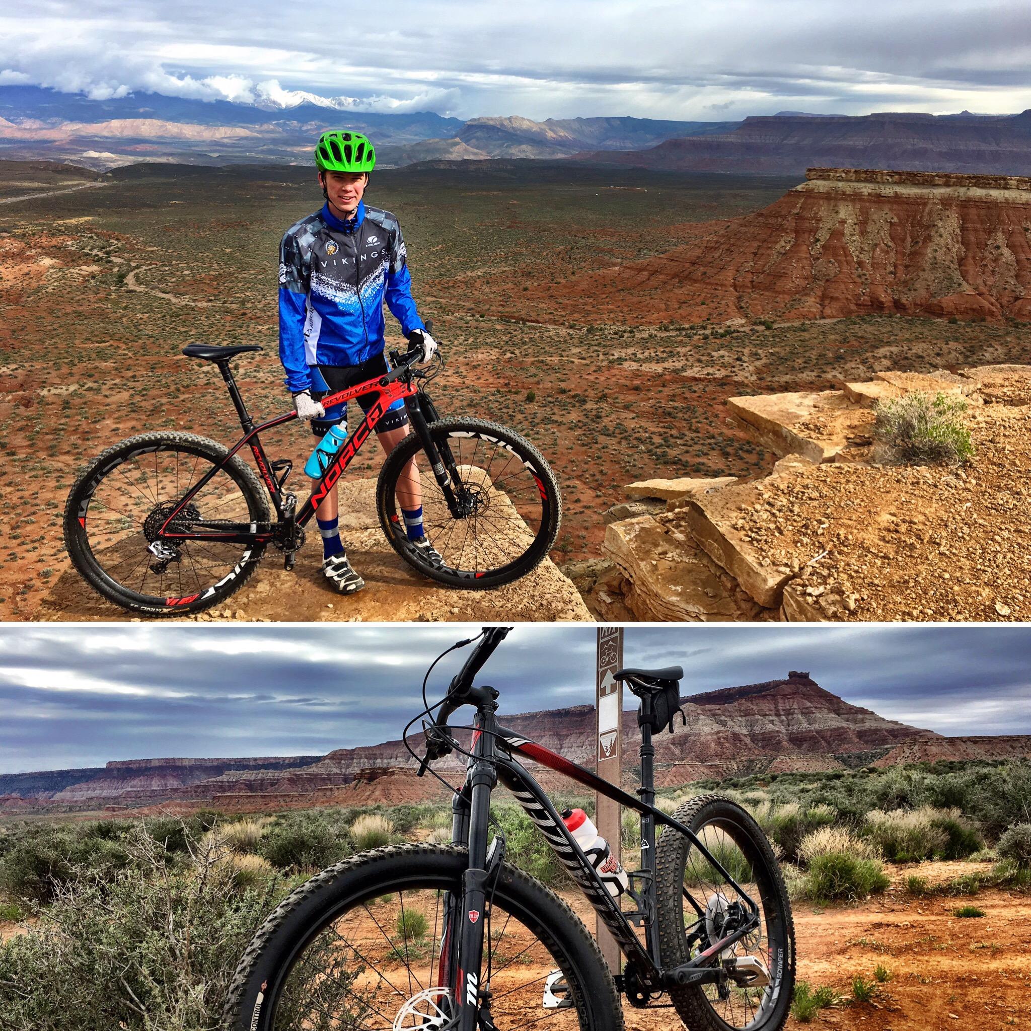 A young cyclist in a blue and black jersey, wearing a bright green helmet, stands next to a mountain bike on a rocky ledge overlooking a vast desert landscape. The scene features red rock formations and distant mountains under a cloudy sky. The image also includes a close-up of the bike, highlighting its design and surrounding vegetation. J.E.M. Trail mountain bike trail.
