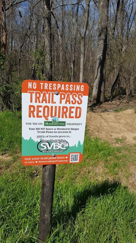 Sign indicating "No Trespassing" and that a "Trail Pass" is required for access to Massanutten property, with details about the necessity of a Western Slope Trail Pass and information about the Shenandoah Valley Bicycle Coalition. The sign is mounted on a wooden post surrounded by grass and trees. Massanutten Western Slope mountain bike trail.