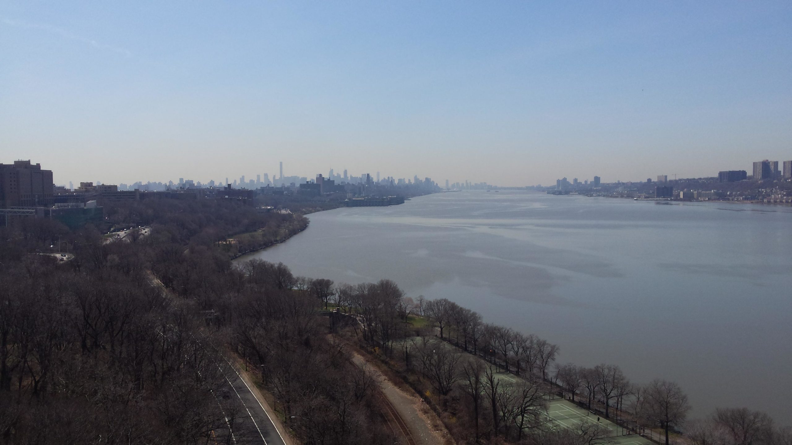 A panoramic view of a river flanked by bare trees and urban buildings, with a skyline in the distance under a clear blue sky. The still water reflects the surroundings, creating a serene atmosphere. West Street Greenway mountain bike trail.