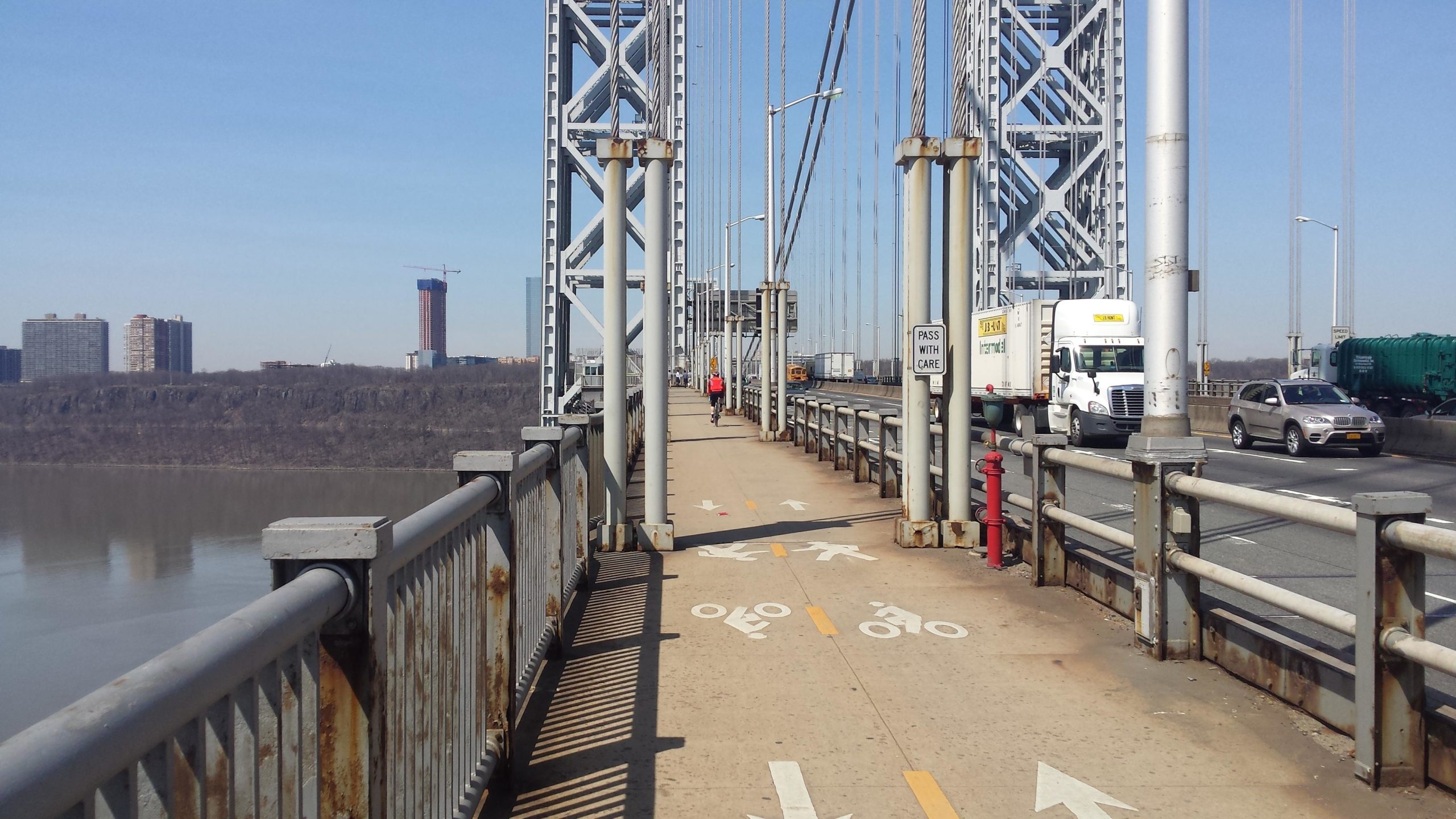 A view of a bridge pathway with marked lanes for pedestrians and bicycles, lined by metal railings and support structures. In the background, vehicles are seen on the roadway, and a construction site can be glimpsed in the distance against a clear blue sky. West Street Greenway mountain bike trail.