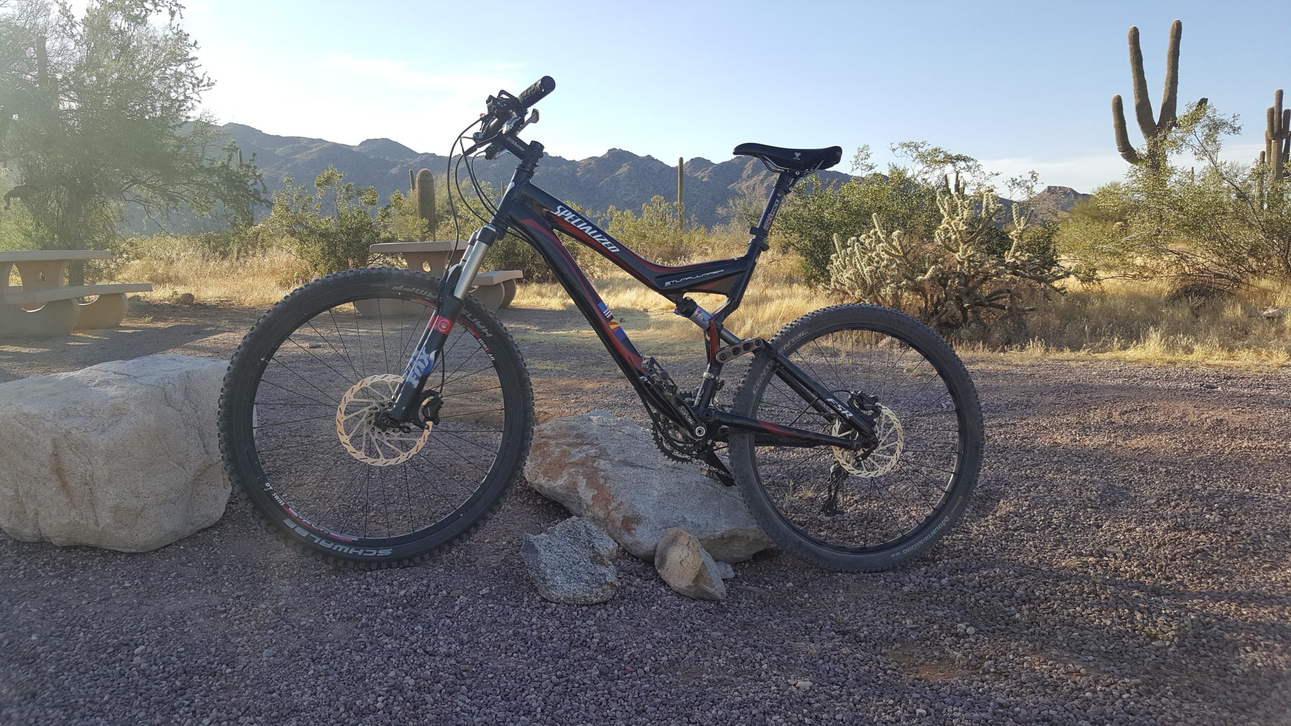 A mountain bike leaning against a rock in a sunlit outdoor setting with desert vegetation and distant mountains in the background. The scene features gravel ground, scattered rocks, and a picnic table in the distance. White Tanks Competitive Track mountain bike trail.