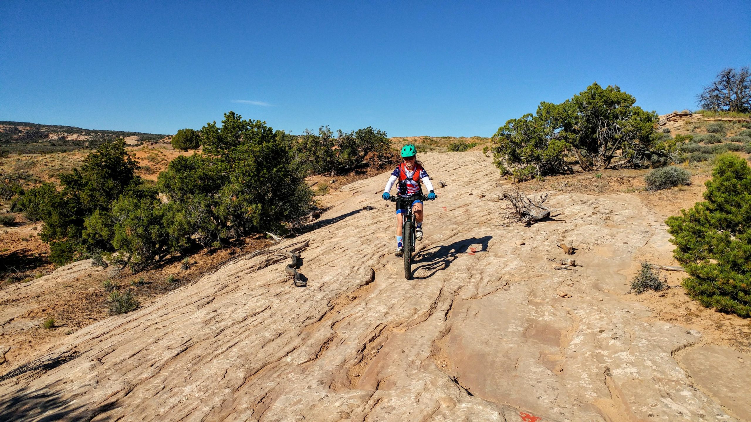 A person riding a mountain bike on a rocky terrain under a clear blue sky, surrounded by sparse vegetation and rolling hills in the background. Navajo Rocks mountain bike trail.