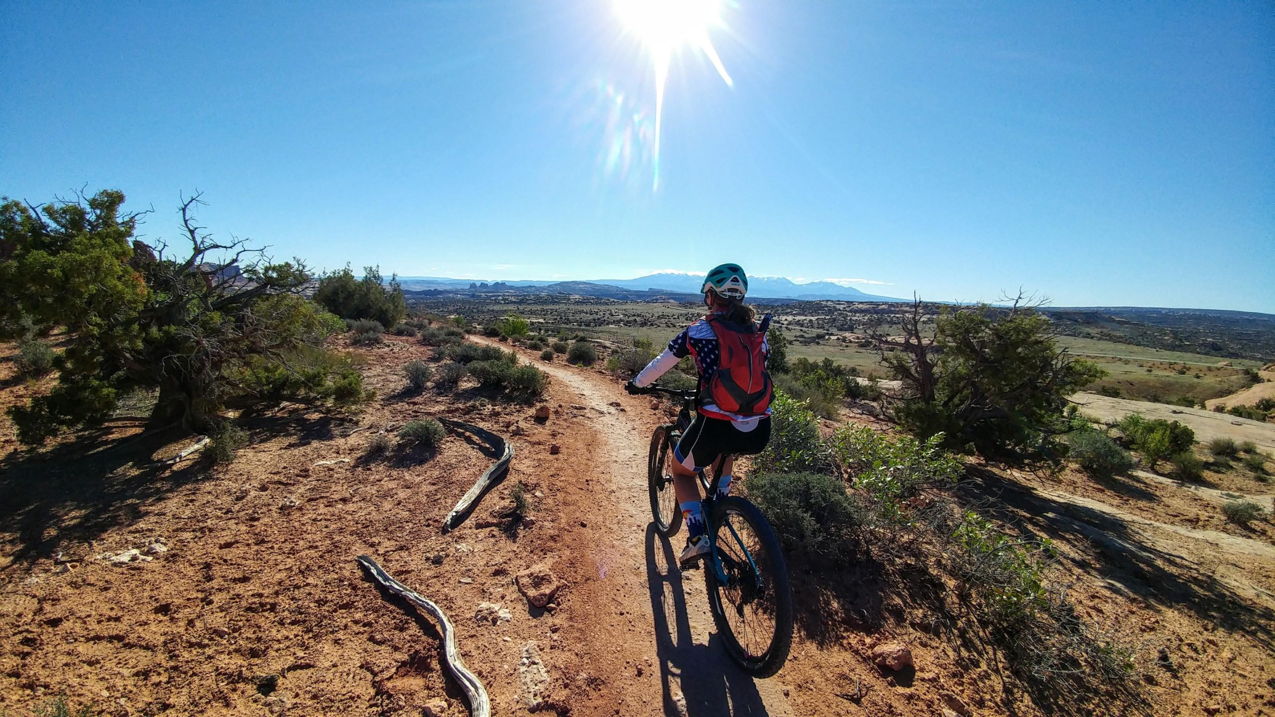 A person riding a mountain bike on a dirt trail surrounded by sparse vegetation, with a bright sun shining overhead and distant mountains visible in the background. Navajo Rocks mountain bike trail.