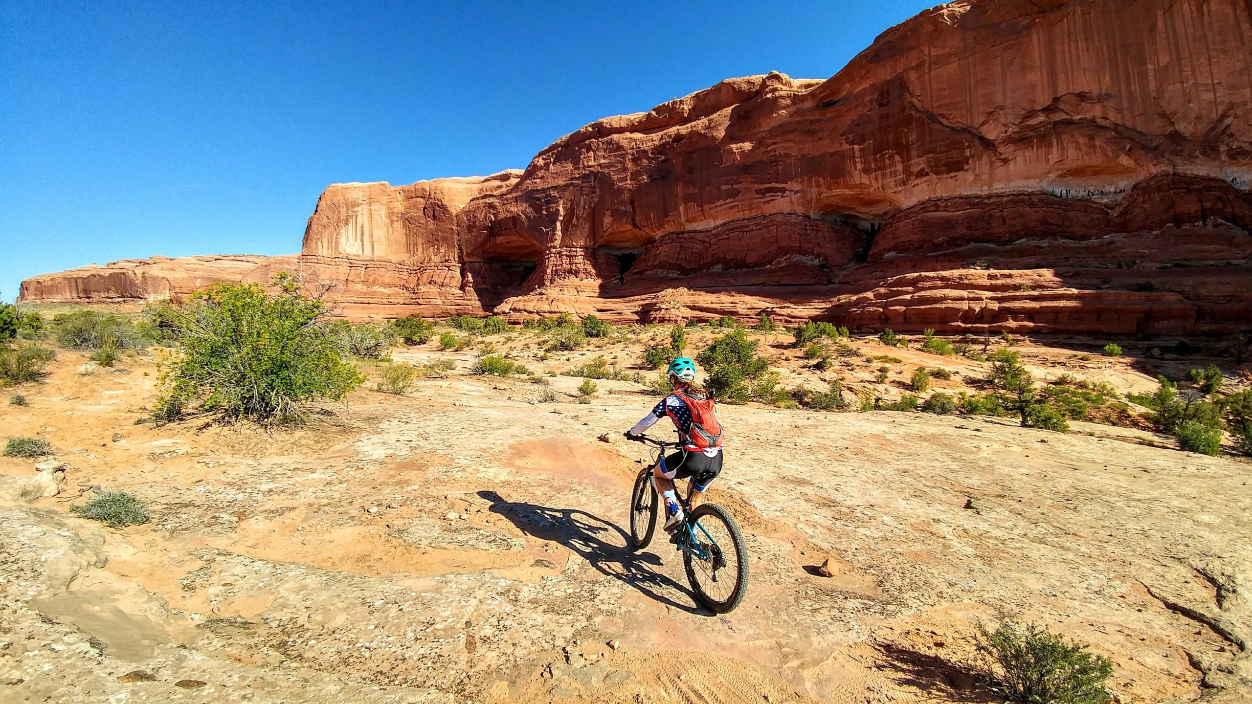 A person riding a mountain bike on a dirt trail surrounded by red rock formations and vegetation under a clear blue sky. Navajo Rocks mountain bike trail.