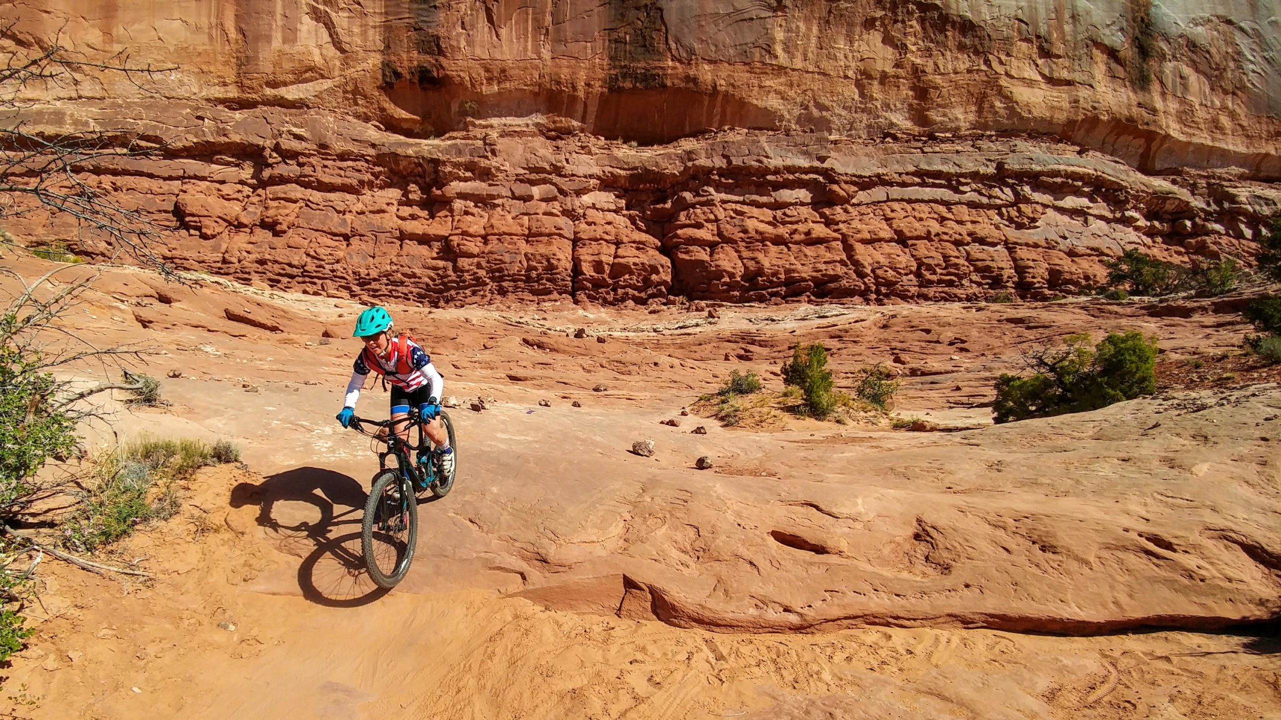 A person riding a mountain bike on a rocky, reddish terrain with steep cliffs in the background. The cyclist is wearing a teal helmet, gloves, and a red and white jersey, navigating through the sunlit landscape. Sparse vegetation is visible along the trail. Navajo Rocks mountain bike trail.