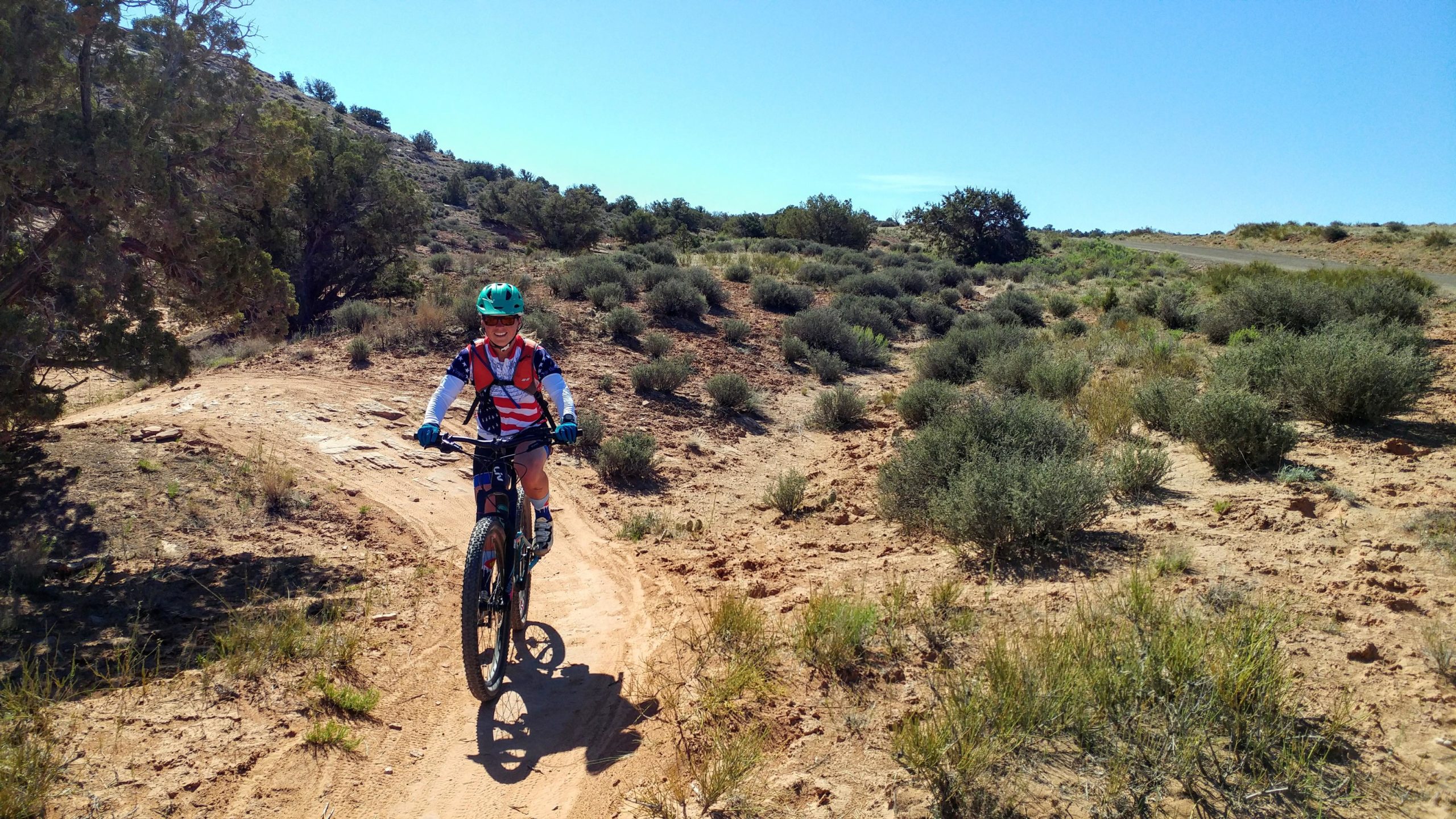 A child riding a mountain bike on a dirt trail in a rocky, arid landscape with sparse vegetation, smiling and wearing a helmet and a patriotic shirt. The background features shrubbery and hills under a clear blue sky. Navajo Rocks mountain bike trail.