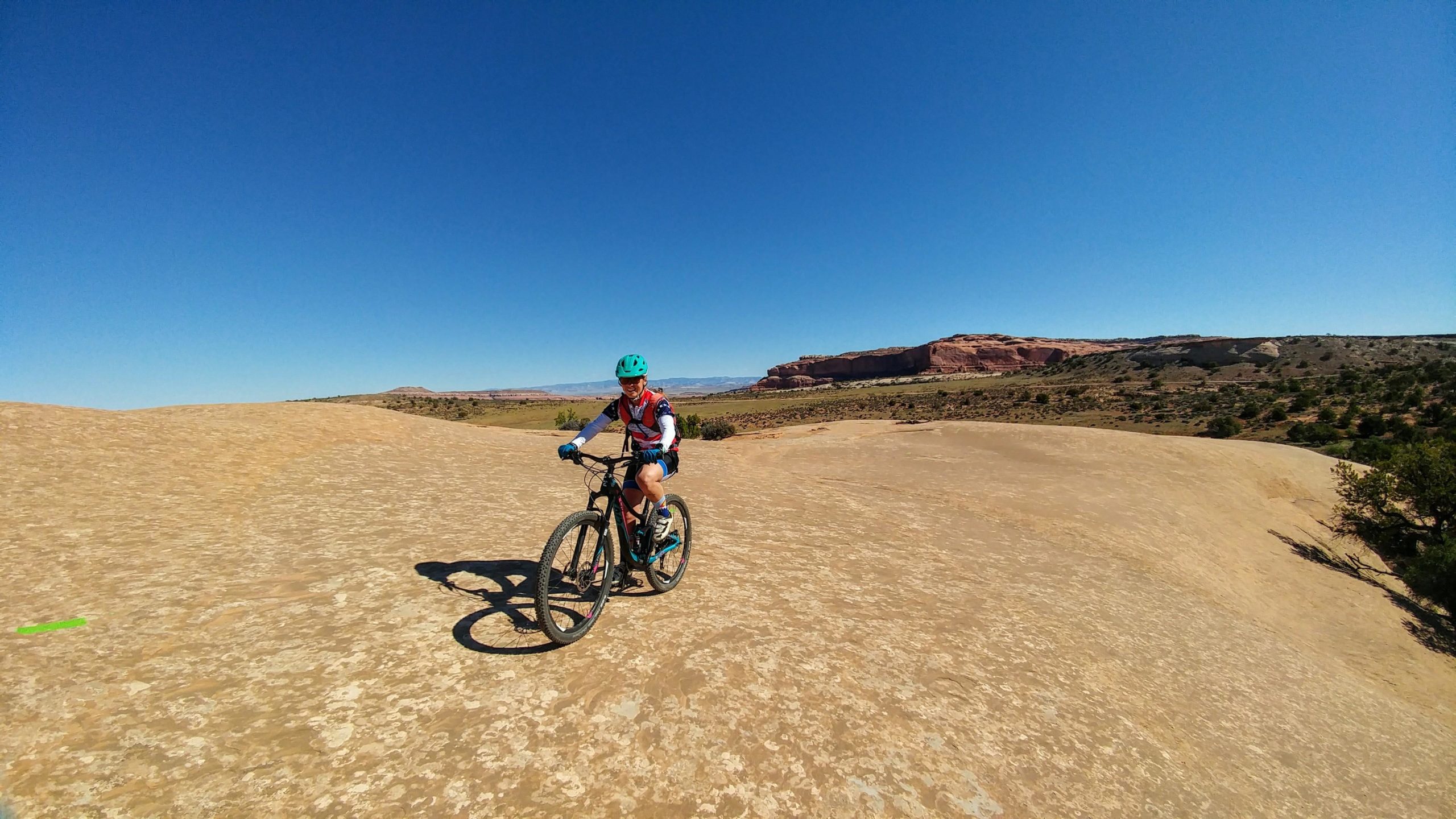 A person riding a mountain bike on a rocky terrain under a clear blue sky, with distant red rock formations and greenery in the background. Navajo Rocks mountain bike trail.