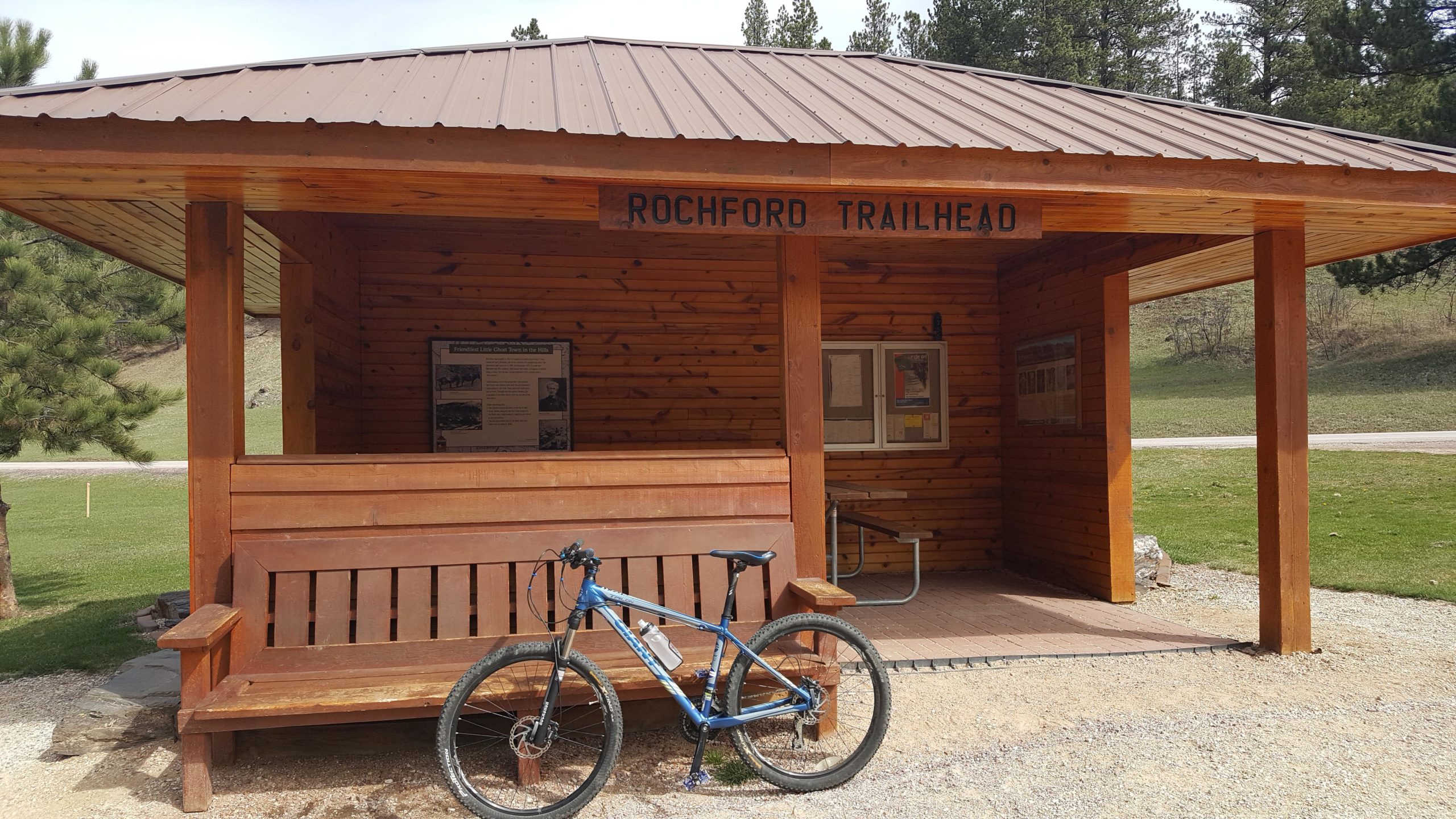 Alt text: Wooden trailhead shelter with a sign reading "Rochford Trailhead," featuring a bench and a blue mountain bike parked beside it. Informational displays are visible inside the shelter, with trees and grassy area in the background. The George S. Mickelson Trail mountain bike trail.