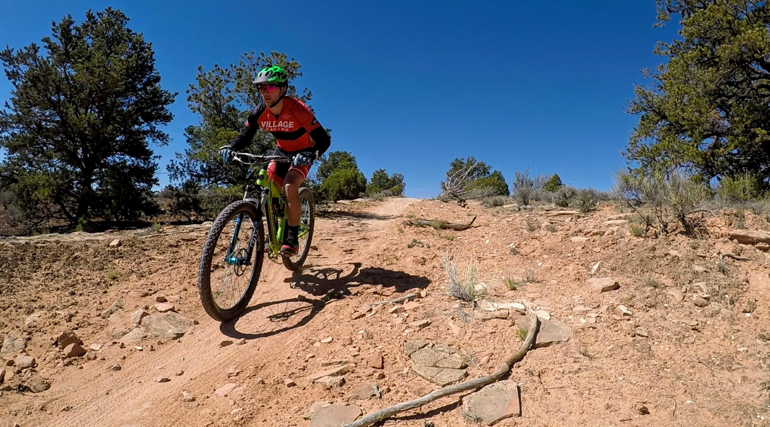 A mountain biker in an orange and black jersey rides on a dirt trail surrounded by trees and a clear blue sky. The bike has a bright green frame, and the biker is actively navigating a hilly section of the terrain. Rocks and scattered vegetation are visible along the path. Navajo Rocks mountain bike trail.