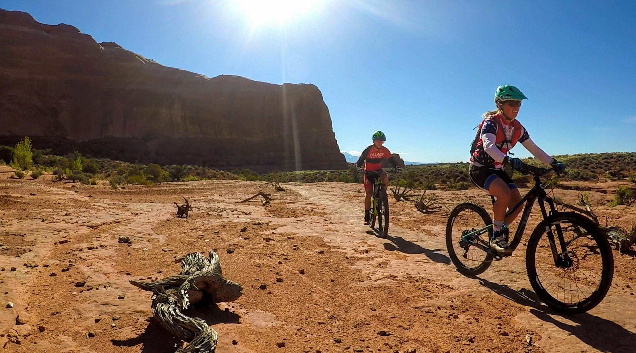 Two mountain bikers ride on a rocky dirt trail in a desert landscape under a bright blue sky. The sun shines overhead, illuminating the stunning rock formations in the background. One rider, wearing a red and black jersey, is navigating the trail, while the other, in a colorful jersey and helmet, smiles as they bike alongside. Sparse vegetation and scattered rocks are visible on the ground, adding to the rugged outdoor ambiance. Navajo Rocks mountain bike trail.