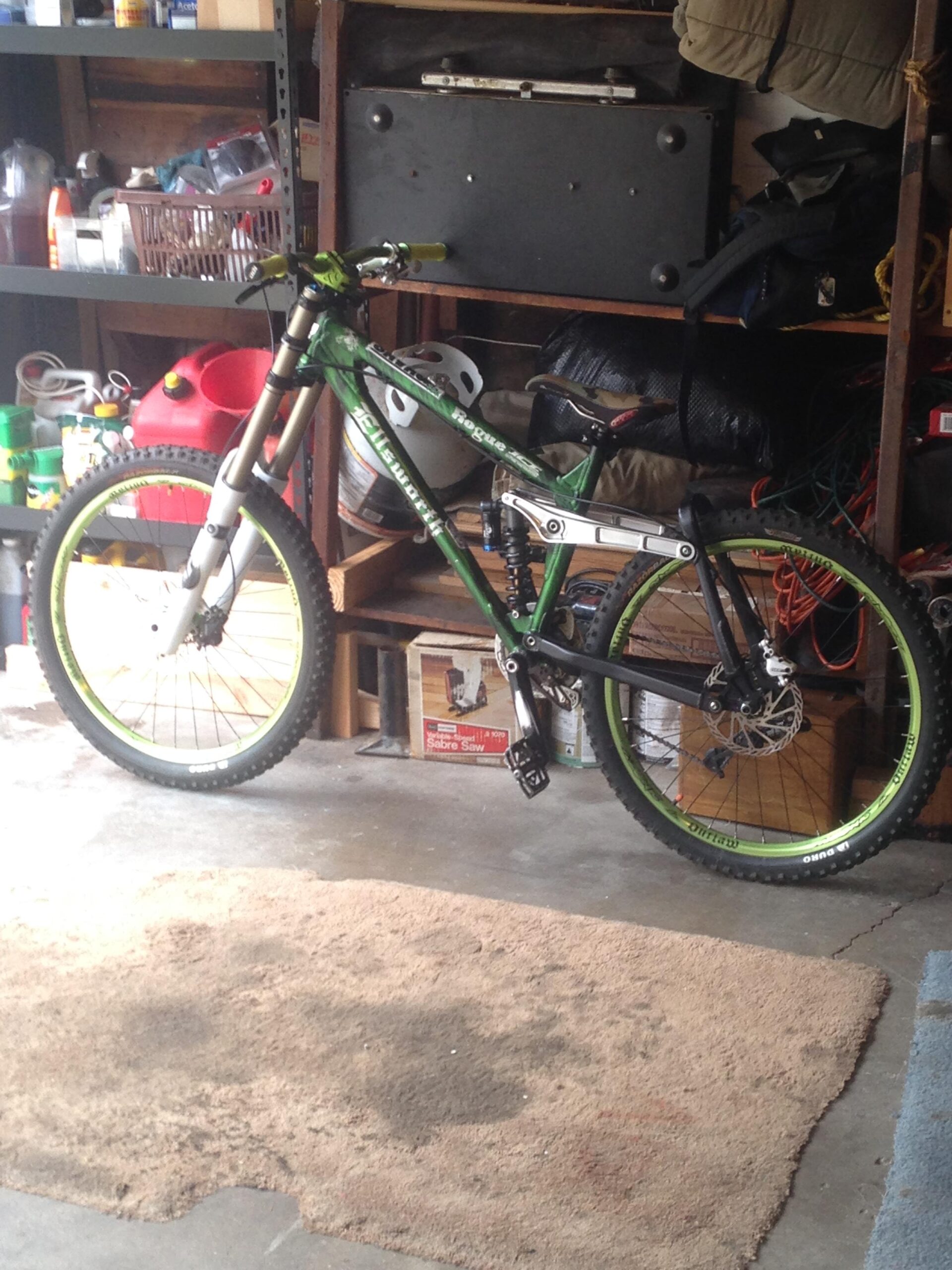 Ellsworth Rogue: A mountain bike with a green and black frame is leaning against a wall in a cluttered garage setting. The bike features wide tires with bright green rims, hydraulic shocks, and a distinct handlebar design. Various items, including a helmet, tools, and storage bins, are visible in the background among the garage clutter. The floor has a beige rug with some stains, and the overall lighting is soft and natural.