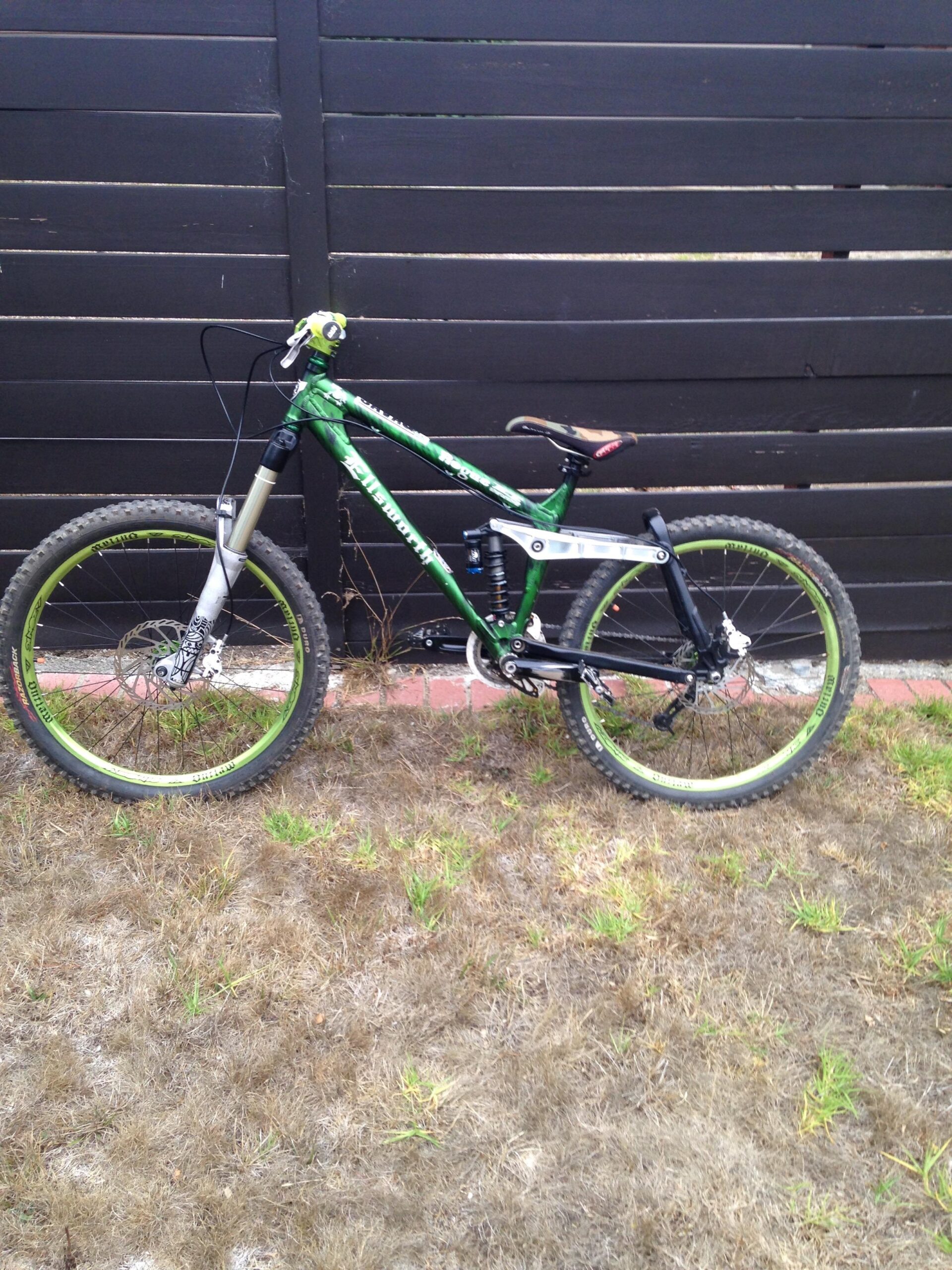 Ellsworth Rogue: A green and black mountain bike is leaning against a dark wooden fence, positioned on a patch of dry grass with some green blades peeking through. The bike features prominent suspension components and distinctively designed wheels.