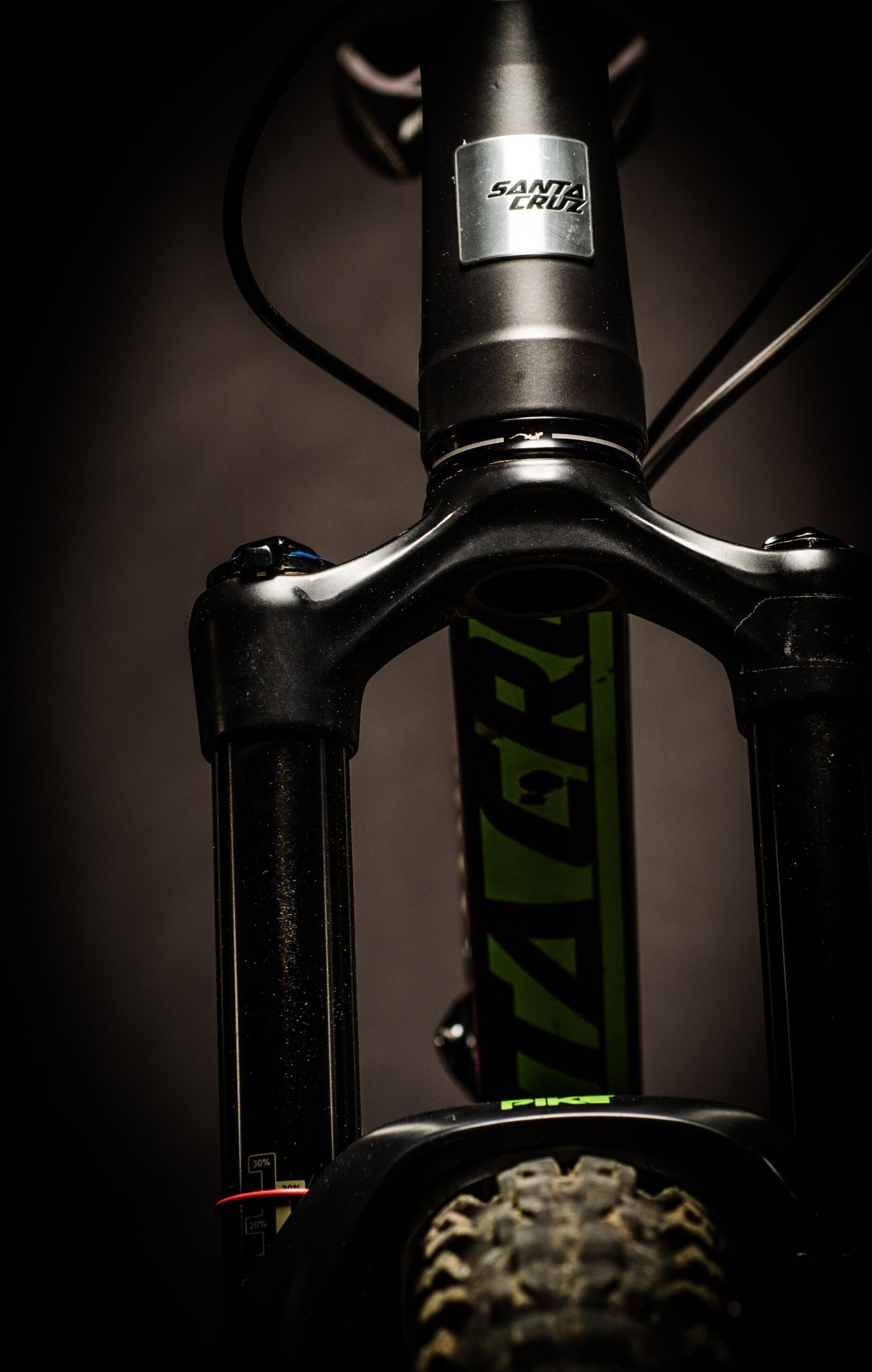 Santa Cruz Bronson: Close-up image of a mountain bike's front suspension fork, featuring a black and green frame with visible branding. The photo emphasizes the fork's details, including the adjustable settings near the axle and the tire tread. The background is dark, enhancing the focus on the bike components.