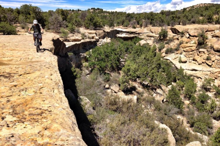 A mountain biker navigates a rocky trail along the edge of a canyon, surrounded by lush greenery and blue skies with scattered clouds. The terrain features a dramatic drop-off on one side, highlighting the thrill of outdoor biking in a natural setting.