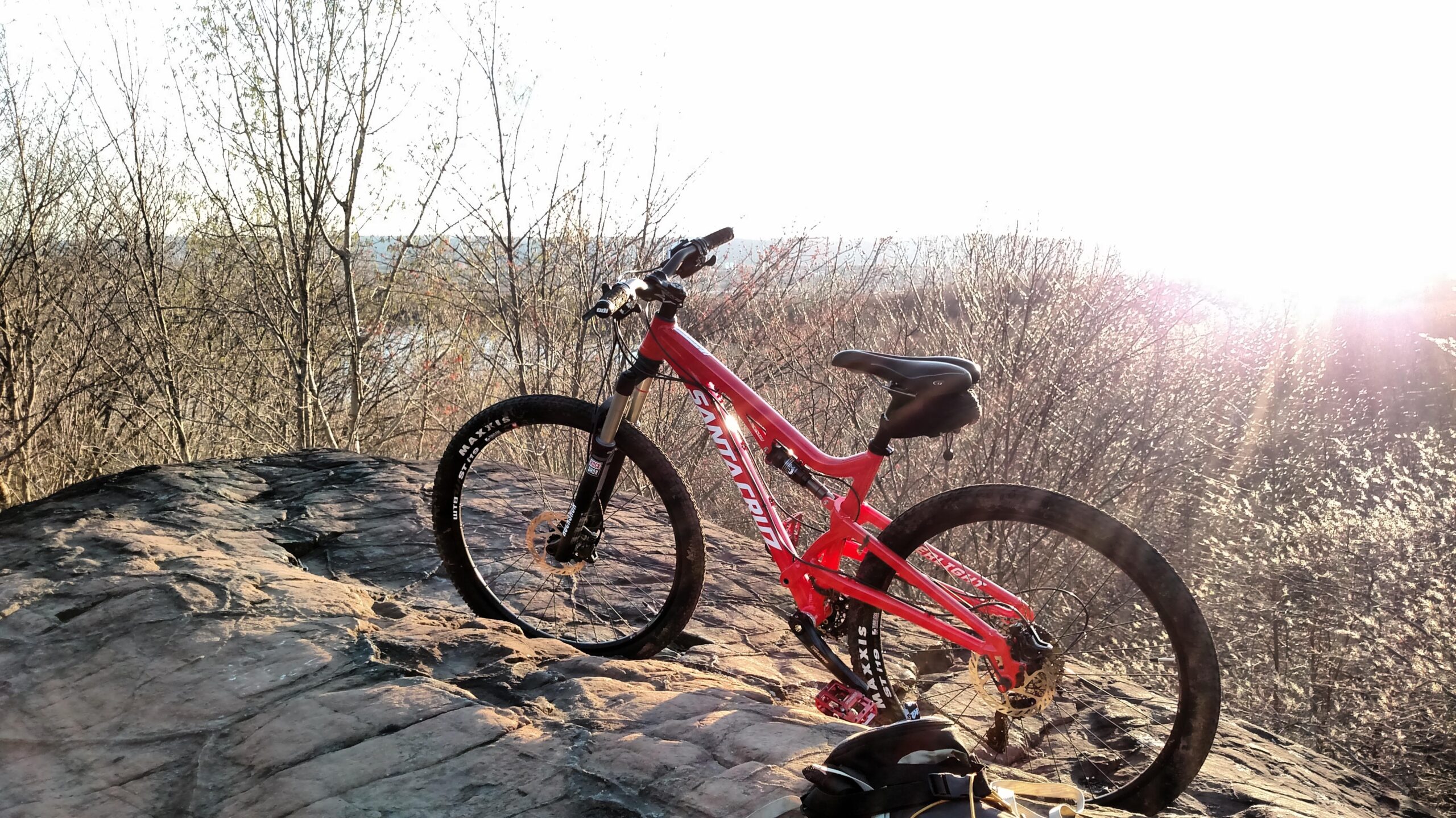 Santa Cruz Superlight: A bright red mountain bike leaning against a rock outcrop, surrounded by bare trees and bathed in golden sunlight, with a scenic view in the background. A small backpack is positioned near the bike.