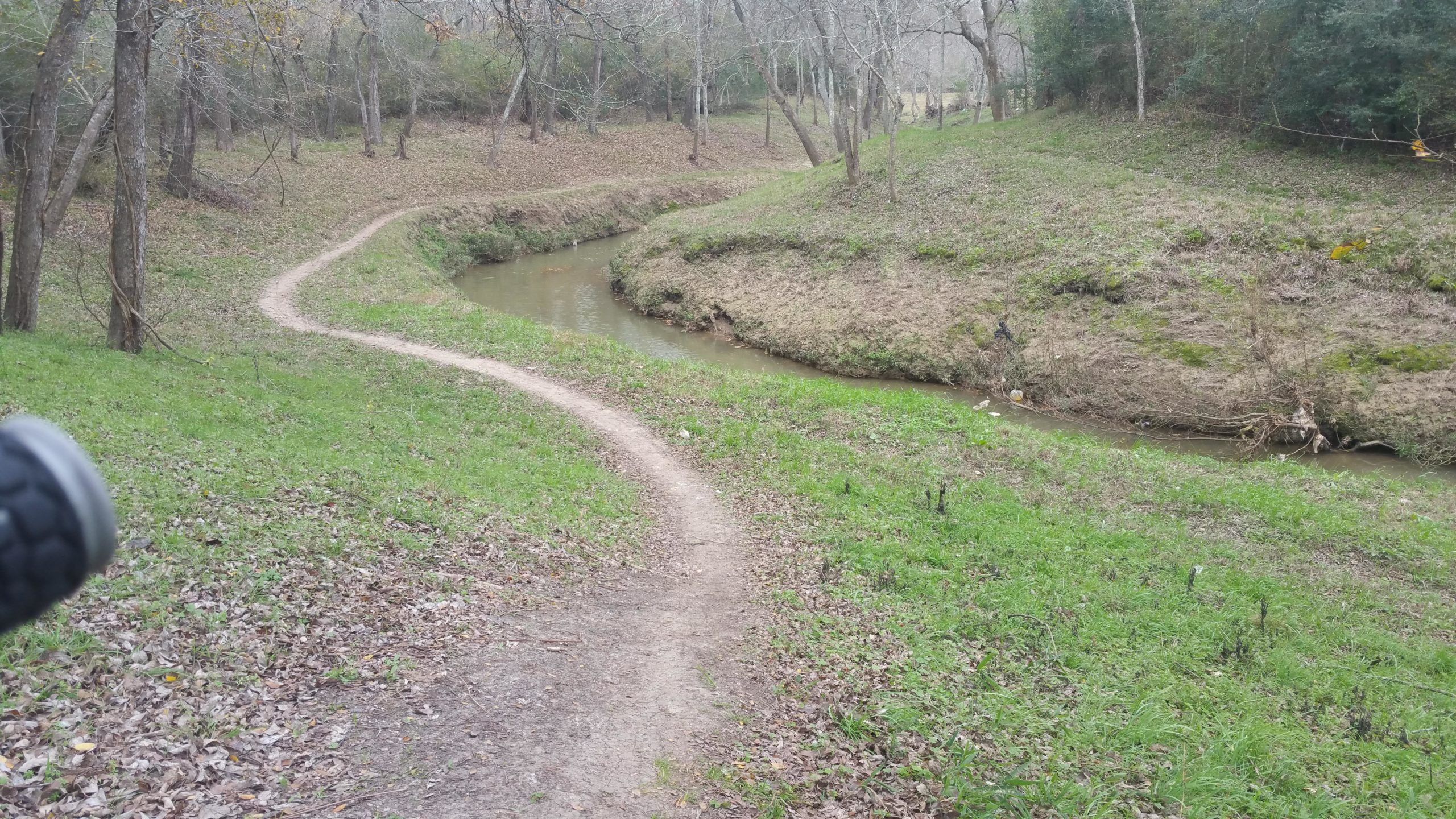 A winding dirt path curves alongside a small, calm stream in a wooded area. Tall grass and scattered leaves line the path, while trees with bare branches provide a natural backdrop. The scene is serene and evokes a sense of tranquility in nature. Armand Bayou Trail mountain bike trail.