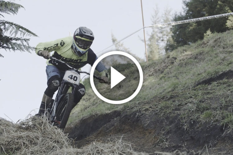 A mountain biker in a bright yellow shirt and blue shorts navigates a downhill track, with trees and grass in the background. The biker is focused and wearing protective gear, including a helmet and goggles, showcasing dynamic movement as they race down the slope. A play button overlay indicates that the image is part of a video.