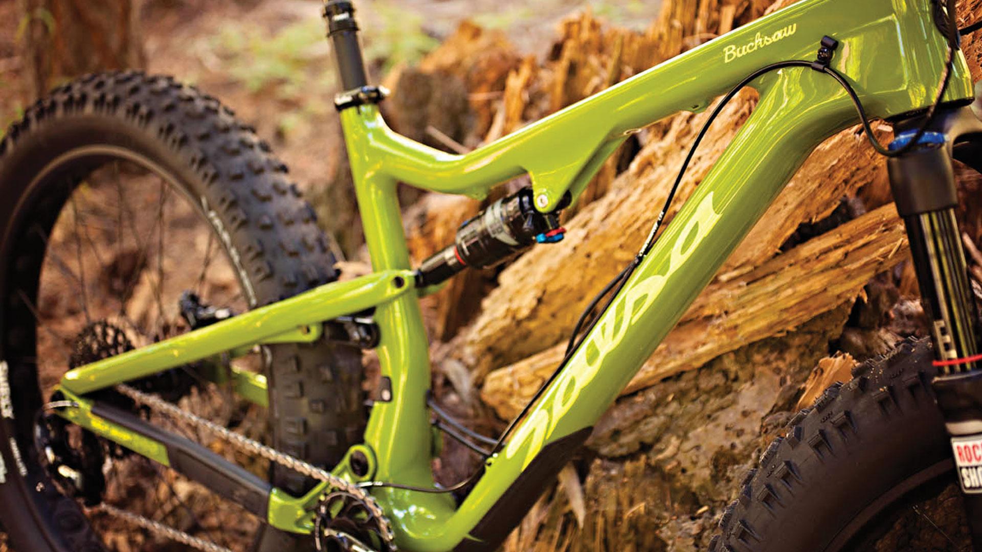Salsa Bucksaw Carbon: A close-up view of a green fat bike frame, featuring a robust design and thick tires, set against a backdrop of natural wood and earth. The bike is highlighted with a prominent brand name, "Bucksaw," and showcases components like a shock absorber and chain.